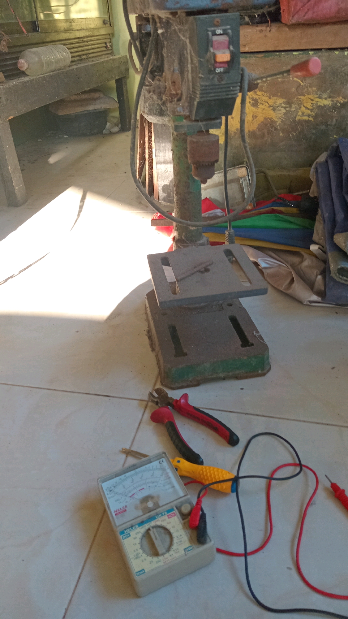 An overhead shot captures a workbench area, likely in Randuagung, Indonesia, where various tools and equipment are gathered on a tiled floor. A drill press, rusted and covered in cobwebs, stands prominently. Its base rests on a green platform, and a metal table with slots is attached to its column. In front of the drill press, a multimeter, a pair of red-handled wire cutters, and a yellow-handled screwdriver are laid out. The scene is illuminated by harsh sunlight casting strong shadows. In the background, a wooden bench supports a plastic water bottle. A collection of colorful fabric items, possibly umbrellas or tarps, are stacked to the right. The overall environment appears to be an indoor workshop or storage area.