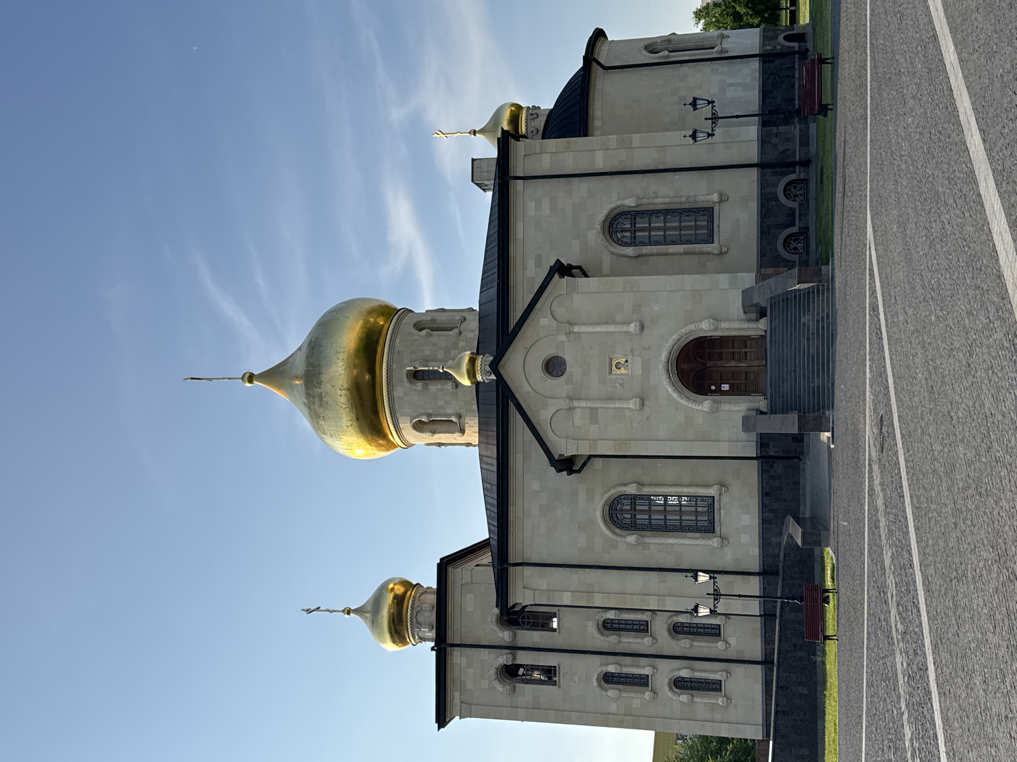 The image shows a large, ornate church with golden domes, located in the city of Kirishi, Russia. The church's facade is made of light-colored stone, with intricate carvings and arched windows. Several golden domes with crosses on top are visible, the largest one dominating the central part of the building. The church is situated in an open plaza paved with cobblestones. Two red benches and lampposts are placed in front of the church. The sky above is clear blue with a few wispy clouds. No people are visible in the image.