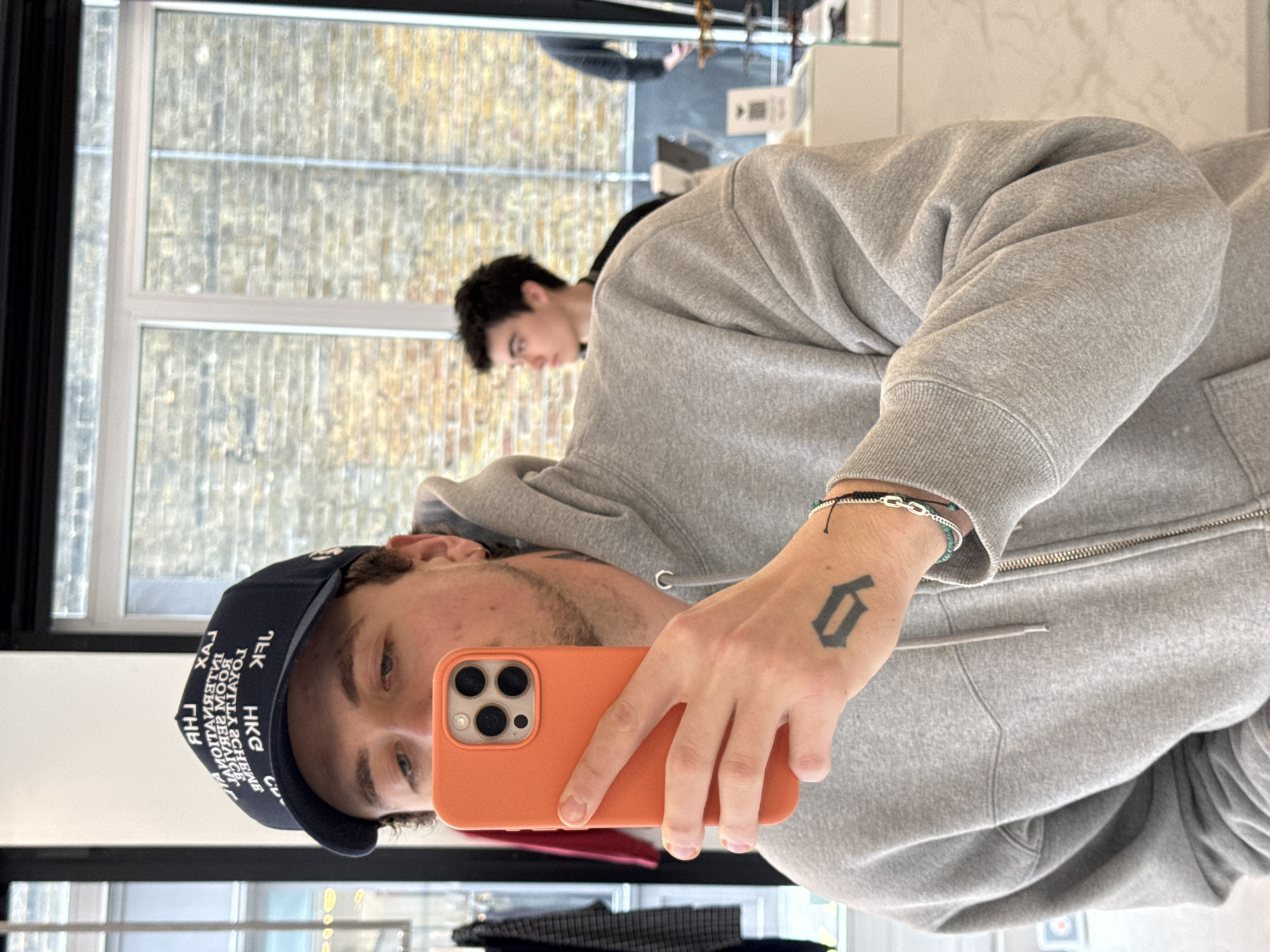 This full-shot image, originally rotated 90 degrees clockwise, captures a mirror selfie taken indoors. The primary subject is a young man, likely in his 20s or early 30s, who is taking the photo. He wears a grey zippered hooded sweatshirt and a dark blue baseball cap worn backward. On his right wrist, he has a silver-colored chain bracelet and another bracelet with small green beads and black string. A distinct tattoo of the letter 'G' in a gothic style is visible on the back of his right hand. He holds an orange-cased smartphone, likely an iPhone, to capture his reflection. His expression is neutral as he looks towards the phone.
In the background, partially obscured, another young man with dark hair is visible, looking downwards or away, suggesting he is also in the same indoor space, perhaps a shop or common area. The setting includes large windows that look out onto a brick building or wall, indicative of an urban environment. The light suggests it's daytime, possibly overcast.
Visible text on the main subject's baseball cap, which appears mirrored due to the reflection, includes 'HKG', 'ROYAL', and other indistinct words in white lettering on the dark blue fabric. The tattoo on his hand clearly shows the letter 'G'. The location is identified as London, United Kingdom, which is consistent with the urban backdrop visible through the windows.