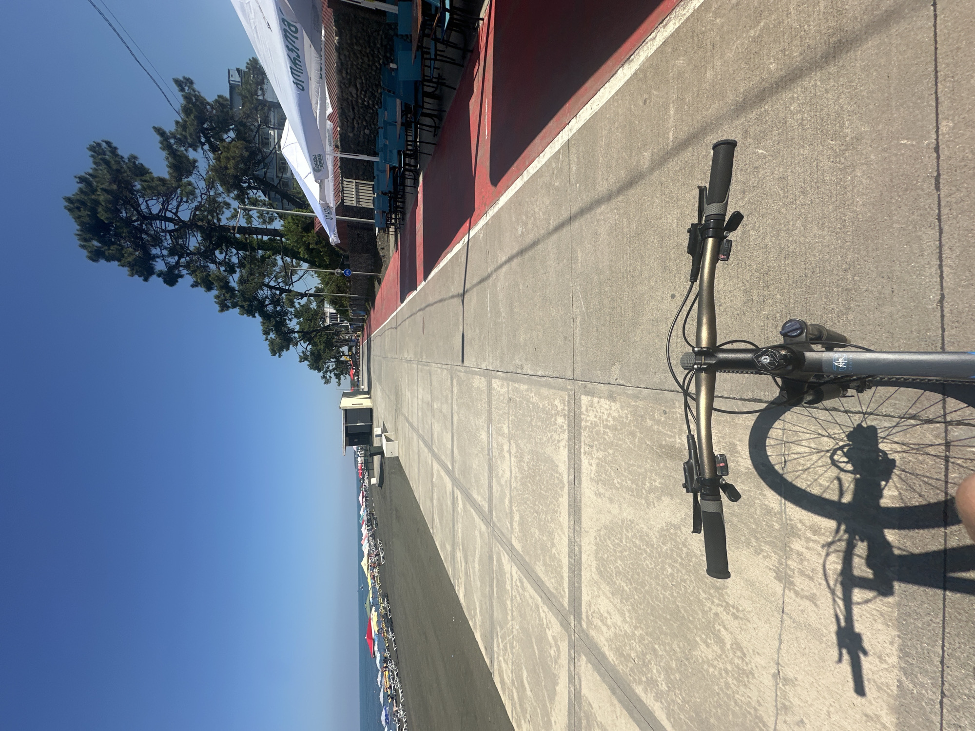 This is a first-person perspective shot of a person riding a bicycle along a paved walkway beside a beach in Ureki, Georgia. The walkway is divided into sections by lines and has a red-painted bike lane adjacent to it. To the left of the walkway is a dark sand beach with numerous rows of lounge chairs and colorful umbrellas. The sea is visible beyond the beach. On the right side of the walkway, there are outdoor seating areas with tables and chairs under white umbrellas, set against a stone wall and trees. The sky is clear and blue. The bicycle's handlebars, front wheel, and a shadow of the bicycle are visible in the foreground.
