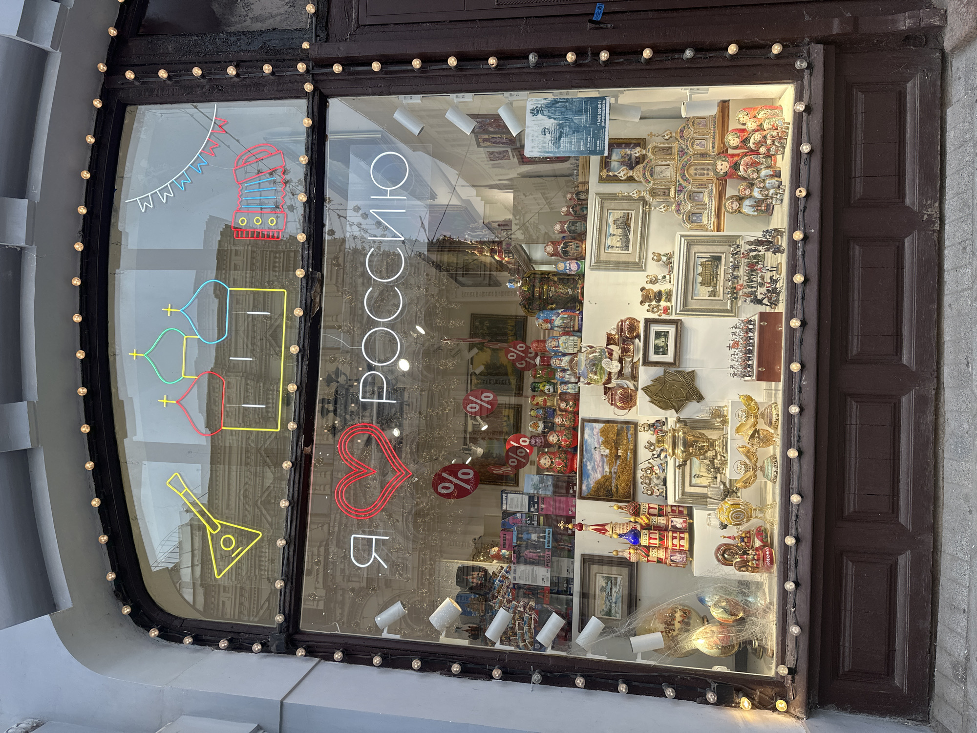 The image shows a shop window display on a street in Moscow, Russia. The window is decorated with neon lights depicting a balalaika, Russian Orthodox church domes, and an accordion, along with a string of small lights. Inside the window, a neon sign reads "Я люблю Россию" which translates to "I love Russia". The display features a variety of Russian souvenirs, including matryoshka dolls, Faberge-style eggs, samovars, small churches, miniature soldiers, and framed artwork. Several discount signs with percentage symbols are also visible. The shop appears to be a souvenir shop selling items representative of Russian culture and heritage. The background reflected in the window shows an urban environment.
