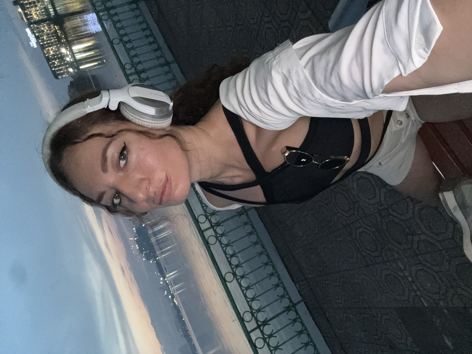 This media file shows a woman wearing white headphones and a white shirt, with a black top underneath. She is wearing sunglasses perched on her chest. The woman is looking at the camera with a neutral expression. The background appears to be a balcony or outdoor seating area with a railing. Beyond the railing, a body of water is visible, reflecting city lights. The sky is a dusky blue, suggesting it is either dawn or dusk. The city lights indicate an urban environment. The image is oriented vertically, with the woman's head on the left side and the background on the right. There is no visible text in the image. The scene suggests a moment of relaxation or contemplation outdoors in a city setting, possibly in Hanoi, Vietnam, given the lighting and reflections.