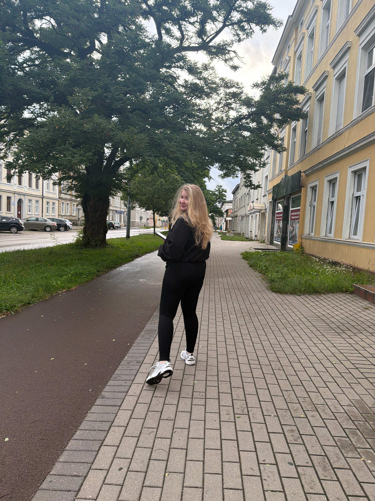 A young blonde woman in black athletic wear and white sneakers walks away from the camera on a cobblestone sidewalk in Koszalin, Poland. She is looking over her shoulder and smiling at the camera. The sidewalk is bordered by a grassy area with small white flowers on the right, and a paved path on the left. Beyond the grassy area, there are buildings with yellow facades and white trim, characteristic of older European architecture. A large tree with lush green foliage stands to the left of the woman, partially obscuring the buildings behind it. Cars are parked along the street in the background. The sky is overcast, suggesting a cloudy day.
