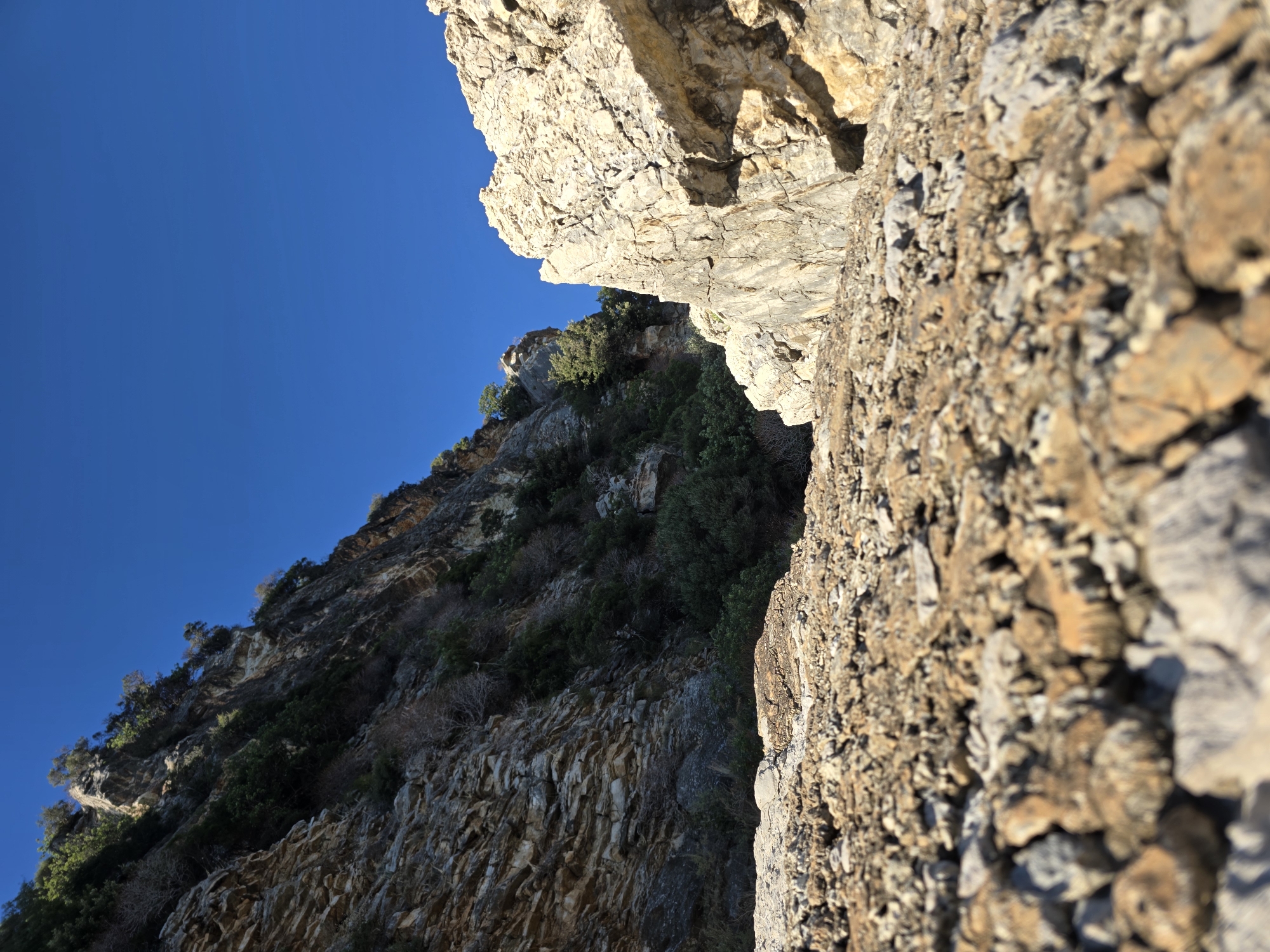 The image depicts a rocky, uneven terrain with steep cliffs on either side, set against a clear, vibrant blue sky. The foreground is dominated by a coarse gravelly surface, with various sizes of stones and rocks, some appearing sharp and jagged, others more rounded. Sunlight illuminates the scene, casting shadows and highlighting the textures of the rocks.
On the left side of the frame, a towering cliff face rises sharply. Its surface is layered and striated, revealing geological formations. Patches of dark green vegetation, including small trees and shrubs, cling to the cliff's surface and crevices, suggesting a rugged, natural environment. The cliff on the right is also prominent, with a lighter, more weathered appearance. It is composed of large, fractured rock masses, creating a sense of imposing scale.
The environment appears arid and mountainous, possibly a coastal cliffside given the proximity to the sea often associated with such formations. The image does not contain any people, animals, or man-made structures, focusing solely on the natural landscape. The overall impression is one of raw, untamed nature, with a sense of geological time evident in the rock formations. The location is identified as Mirište, Montenegro, which is known for its coastal cliffs and rugged terrain.
