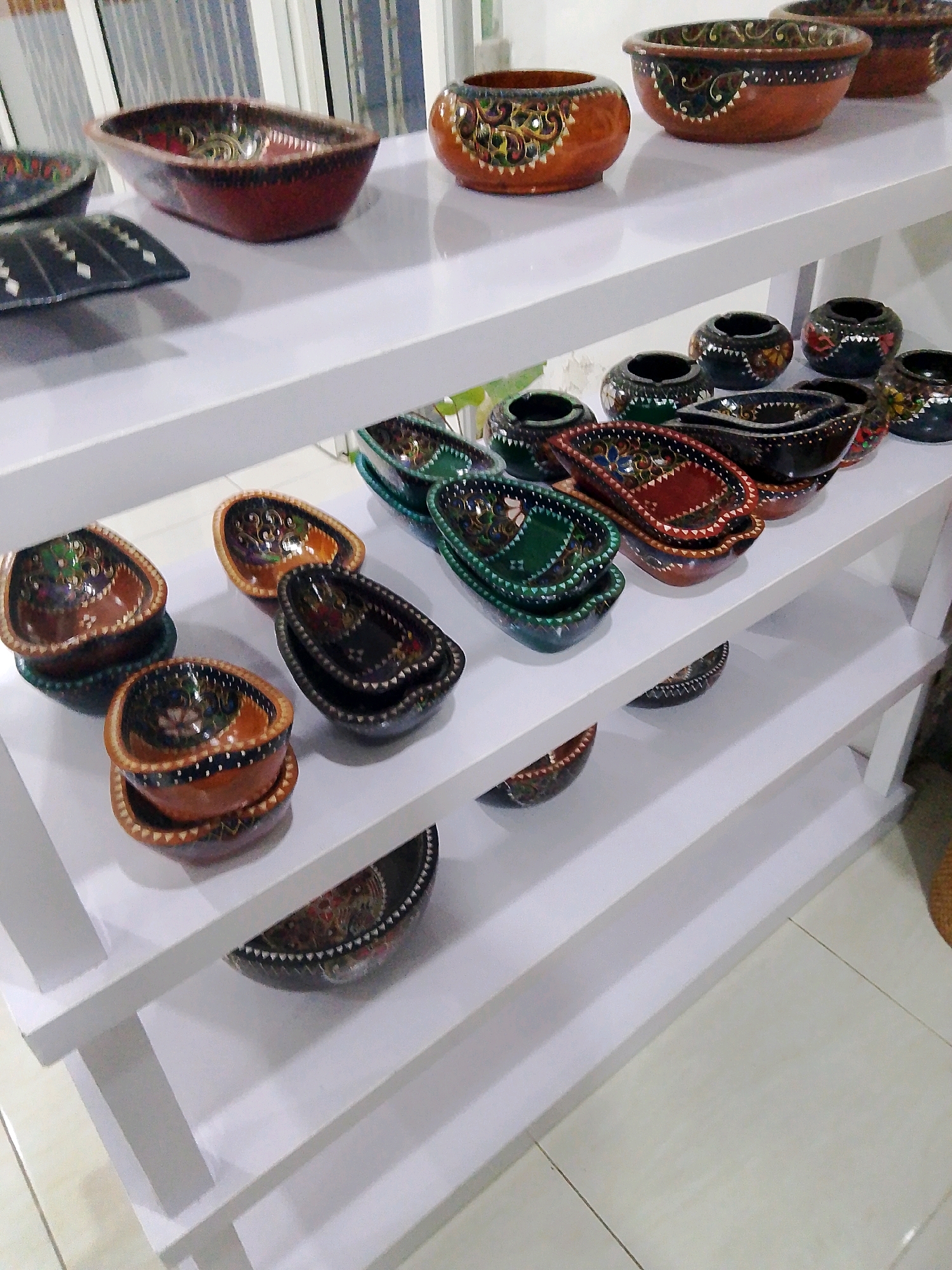 In Bunkate, Indonesia, a white, multi-tiered shelf displays an assortment of decorative ceramic bowls and containers. The top shelf features a few larger, round bowls with intricate painted patterns in shades of orange, brown, and green, one of which has a distinctive floral design with what appear to be small mirrored accents. To the left of these, a shallow, reddish-brown oval bowl with a dark rim showcases a similar decorative style. Further left, a dark, rectangular dish with a geometric white pattern rests on the shelf.
The second shelf from the top holds a collection of smaller, dark-colored bowls, some appearing to be ashtrays, with painted designs in various colors, including green, red, and yellow. There are also several rectangular, boat-shaped bowls in dark brown and green, each with detailed geometric and floral motifs.
The third shelf displays a row of small, colorful, boat-shaped bowls, some appearing to be nested. These bowls are predominantly brown and green, with painted patterns that include swirling lines and diamond shapes. A dark-colored bowl with a serrated rim is positioned to the right of these.
The bottom shelf is less populated, with a dark, patterned bowl partially visible on the left and another decorative bowl in the center. The overall impression is that of a retail display of handcrafted ceramics. The background is indistinct, with hints of vertical blinds or window coverings and a tiled floor.