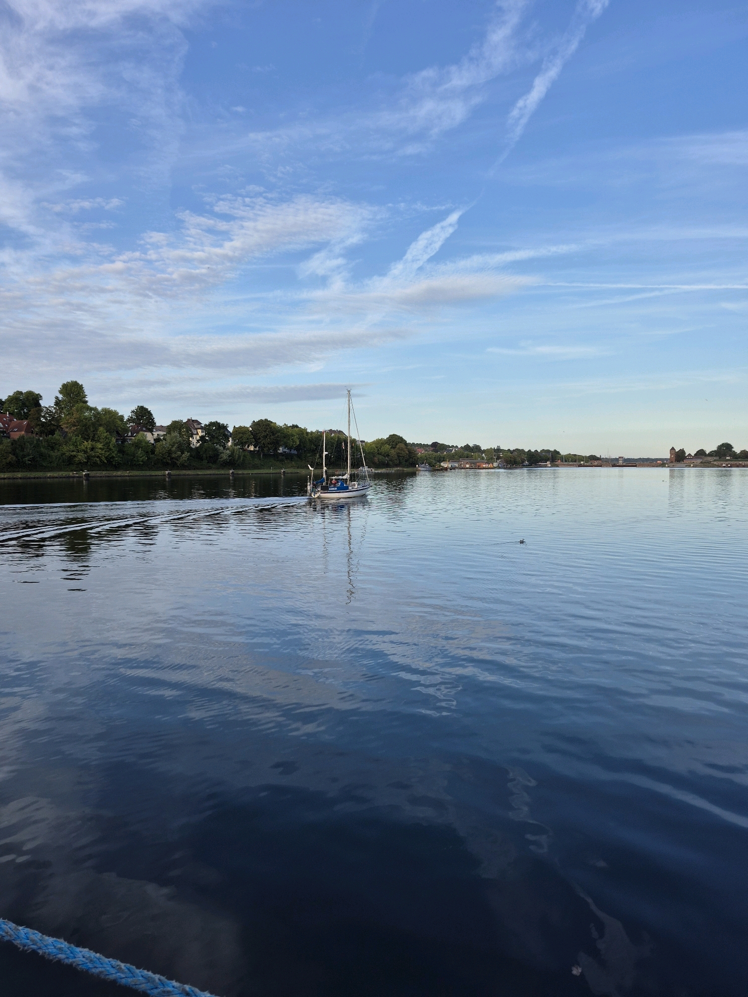 The image depicts a wide, calm body of water with a clear blue sky above, dotted with wispy white clouds. In the mid-ground, a white sailboat with a single mast is sailing away from the viewer, leaving a V-shaped wake on the water. The shoreline in the background is lined with lush green trees and houses with red-tiled roofs, suggesting a residential area. Further in the distance, a red brick structure, possibly a tower or building, can be seen on the right side of the frame. The water's surface reflects the sky and the surrounding environment, creating a serene and tranquil atmosphere. A blue rope is visible in the foreground, hinting that the photo was taken from a boat or pier. The overall scene is characteristic of a waterfront in Kiel, Germany, known for its maritime activities and picturesque landscapes.