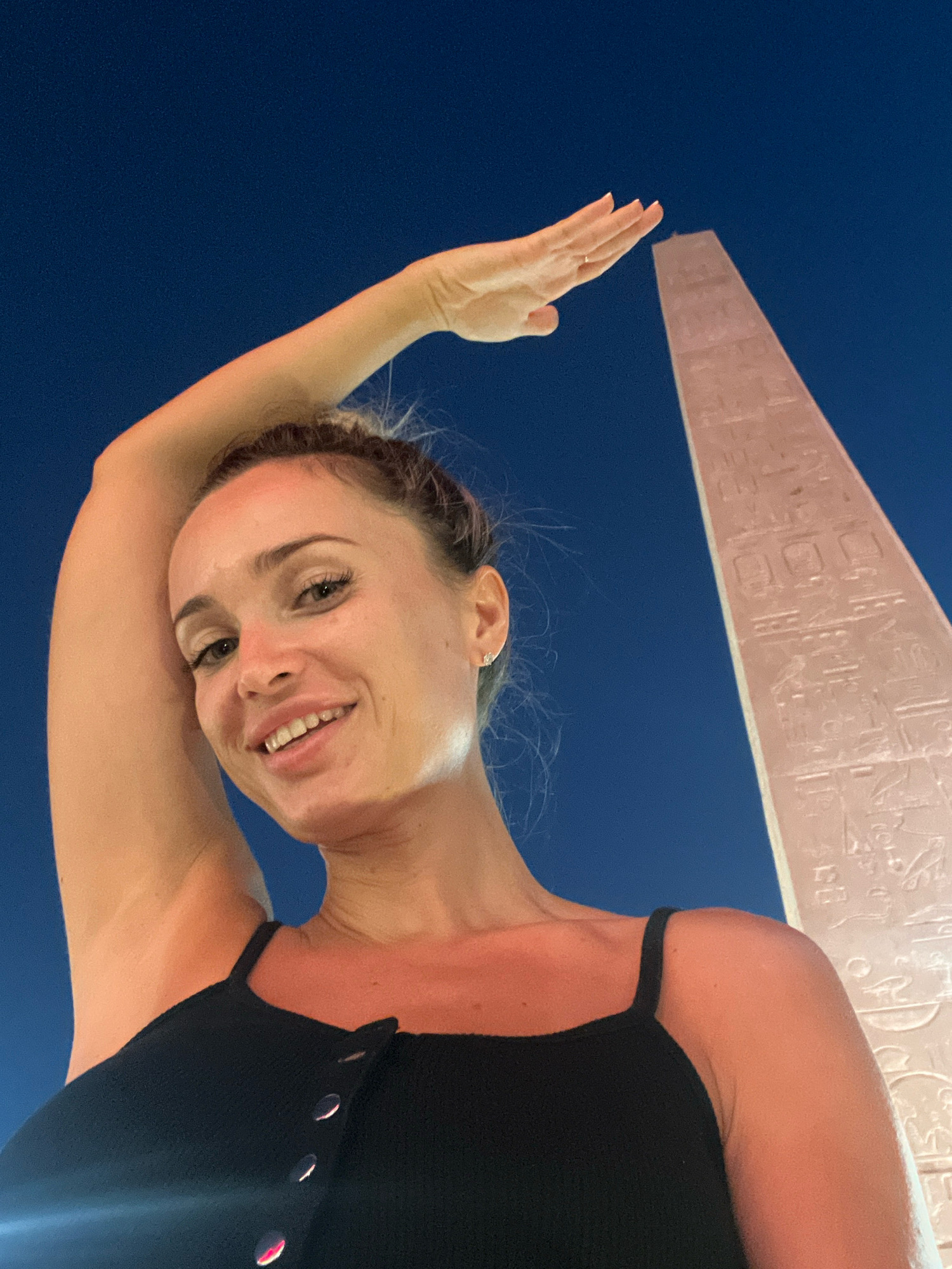 A young woman with blonde hair in a bun smiles at the camera, her right arm raised above her head with her hand outstretched towards a tall, ancient Egyptian obelisk. The obelisk is covered in hieroglyphics and is lit by an artificial light source. The background is a dark, clear night sky. The woman is wearing a black tank top. The image appears to be taken at night, possibly in an outdoor tourist area. The location is the Cleopatra Kingdom amusement park in Sharm El Sheikh, Egypt, which features a replica of an obelisk.