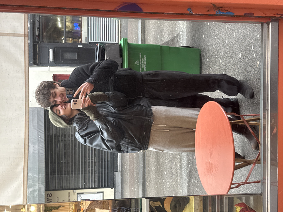 A young adult couple is captured in a mirror selfie on a Parisian street. The female, wearing an olive-green beret, dark leather jacket, olive-green fingerless gloves, and light grey sweatpants, holds a light-colored smartphone with both hands, positioned to capture their reflection. She is leaning her head against the male's cheek, smiling faintly. The male, with curly dark hair, wears a dark sherpa-lined jacket, dark sweatpants, and dark shoes; his arm is around the female, and he is also holding a dark-colored smartphone, partially visible.
The scene reflects a wet street with grey pavement. To the left, a storefront labeled "S'M BOULANGERIE" (reversed) displays baked goods. To the right, another building features a dark storefront with a metal security shutter. A prominent, bright green public recycling bin with white French text ("TRIER C'EST" visible) stands behind the male. In the immediate foreground, a round, reddish-orange metal bistro table is partially visible at the bottom left. The image is framed by an orange border on the right, indicating the edge of the reflective surface, likely a shop window or mirror, in the city of Paris, France.