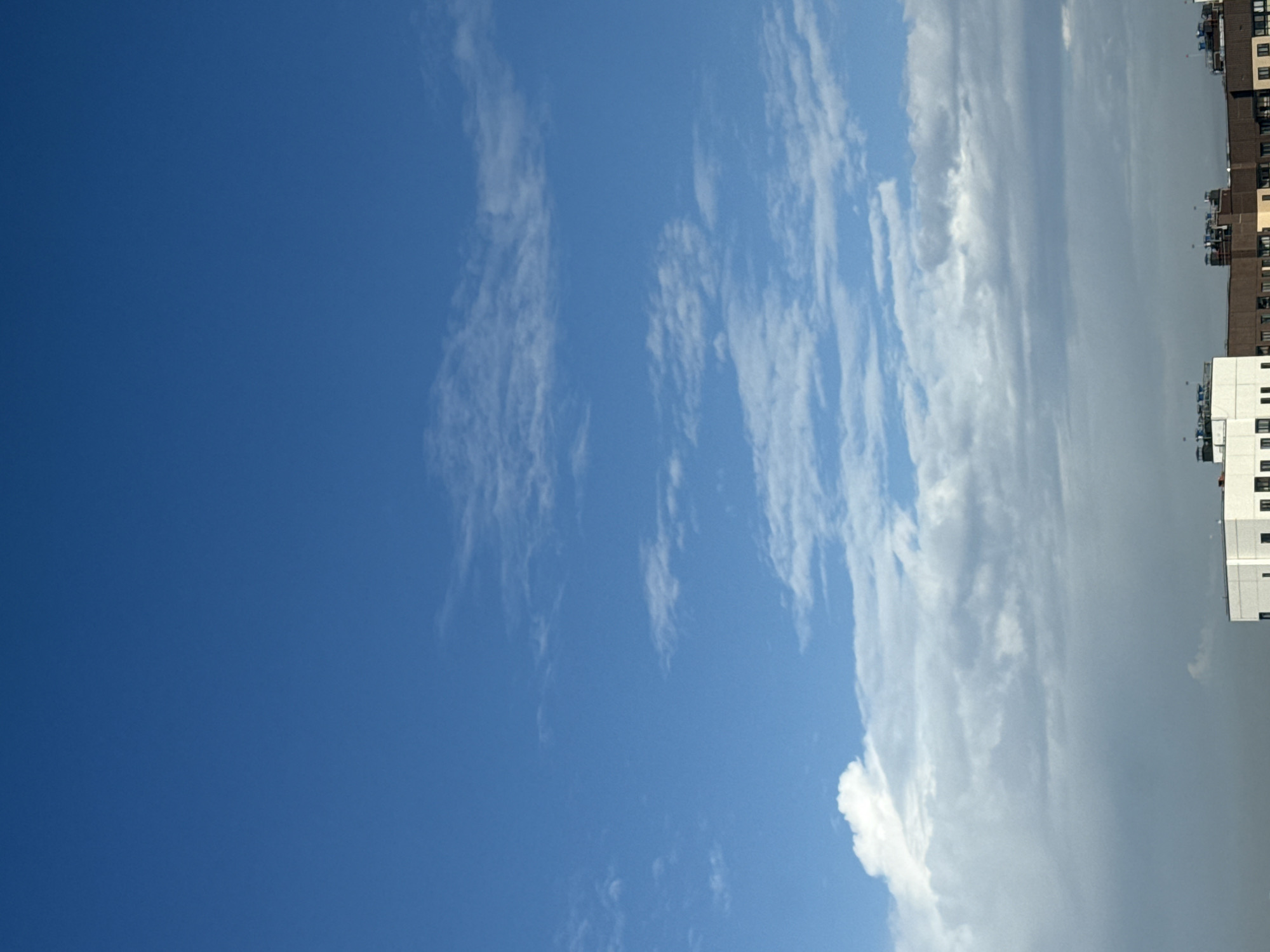 The image presents a view of the sky in what appears to be an urban area. A clear, deep blue sky occupies a significant portion of the left side of the frame, gradually transitioning into a sky with wispy, elongated clouds towards the right. Further to the right, the cloud cover becomes denser, taking on a greyish-white hue. The upper-right edge of the image shows buildings of varying heights and colors, indicative of an urban environment. While specific architectural details that would pinpoint the exact location in Lyubertsy, Russia are not discernible, the presence of multi-story buildings and the general urban sky conditions suggest a built-up environment.