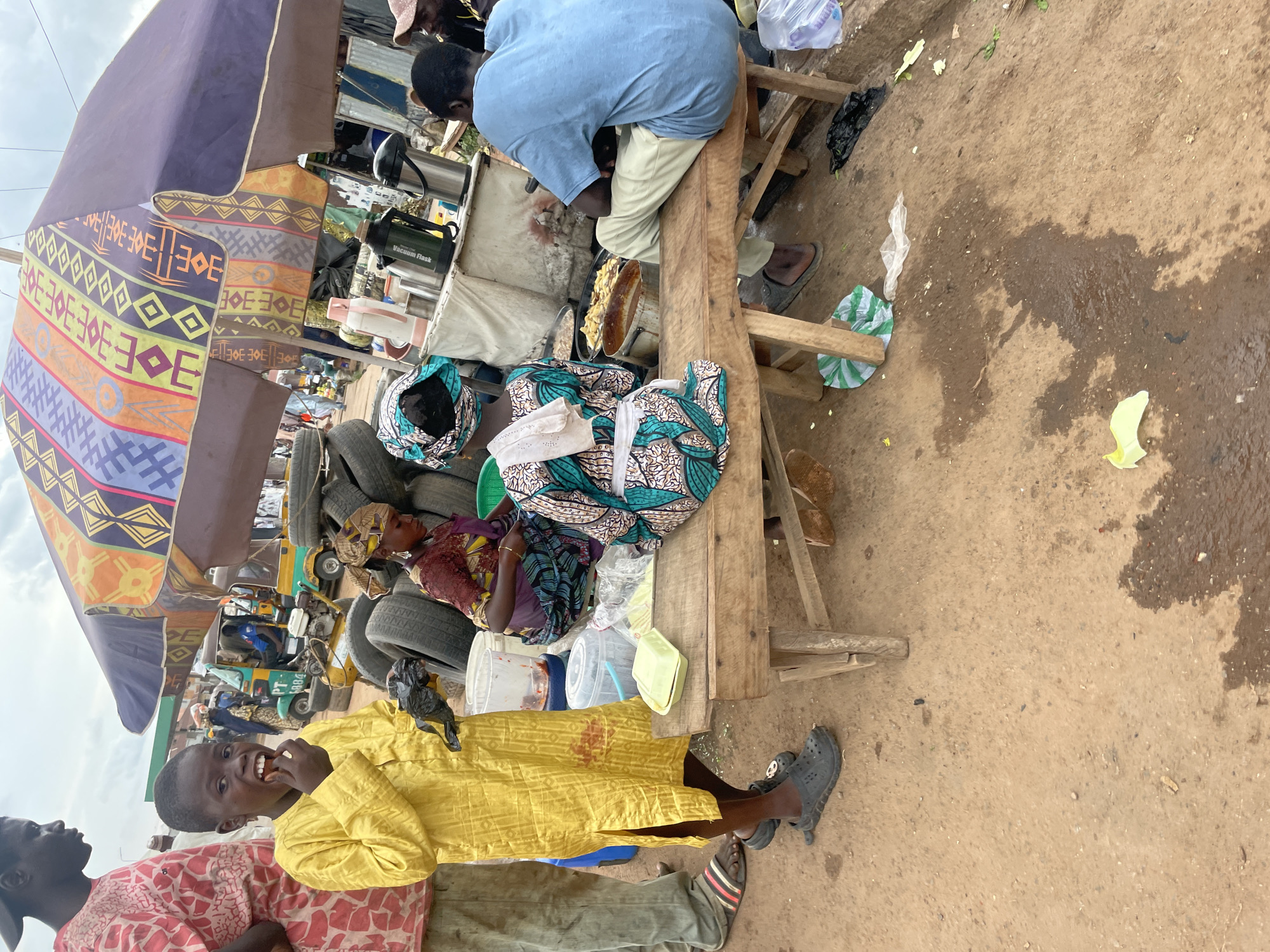 A boy in a yellow tunic eats from his hand in front of a table laden with food. He is standing on a dirt ground, next to a man in a patterned shirt and gray trousers. Behind them, a large, colorful umbrella shades a food stall. Several women are visible, some tending to the food, and others sitting or standing around. One woman, wearing a patterned headscarf and a blue and green patterned dress, is seated on a wooden bench, as is a man in a blue t-shirt and light-colored pants. The background shows stacks of tires, a tricycle, and other market stalls, suggesting a bustling marketplace scene in Jos, Nigeria. The ground is wet in places, and there is some scattered debris.
