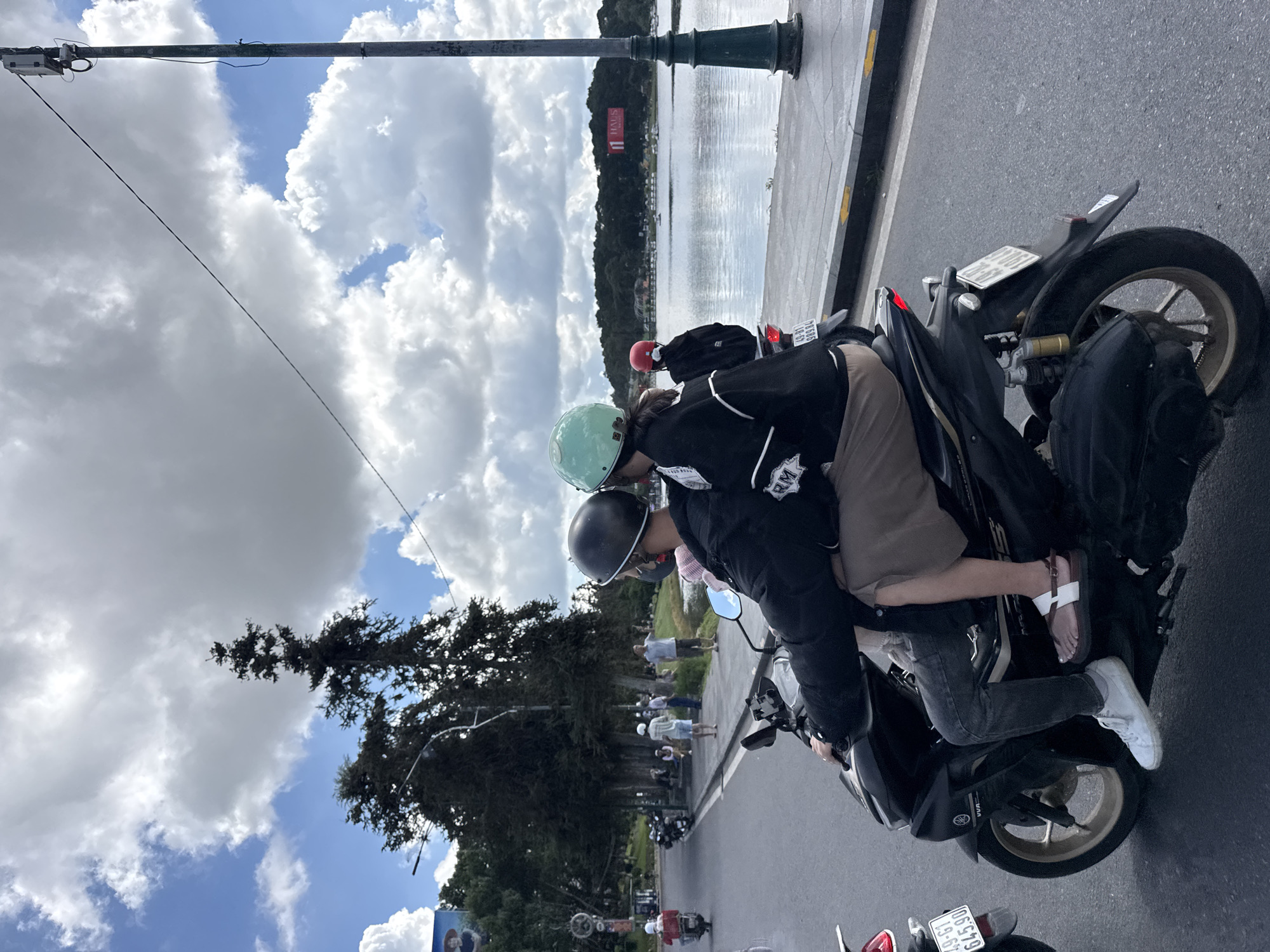 A couple rides a motorcycle along Xuan Huong Lake in Da Lat, Vietnam. The man, in front, wears a black jacket and grey jeans, with a black helmet and a grey face mask. The woman, riding behind, has on a black jacket and a beige skirt, and sports a light green helmet. The motorcycle is black with gold accents. In the background, large trees line the road, with Xuan Huong Lake to the right. A body of water reflects the cloudy blue sky. A lamppost stands tall on the right side of the road, and a billboard displaying "HAUS" is visible across the lake. Other people and motorcycles are present on the road and sidewalk.