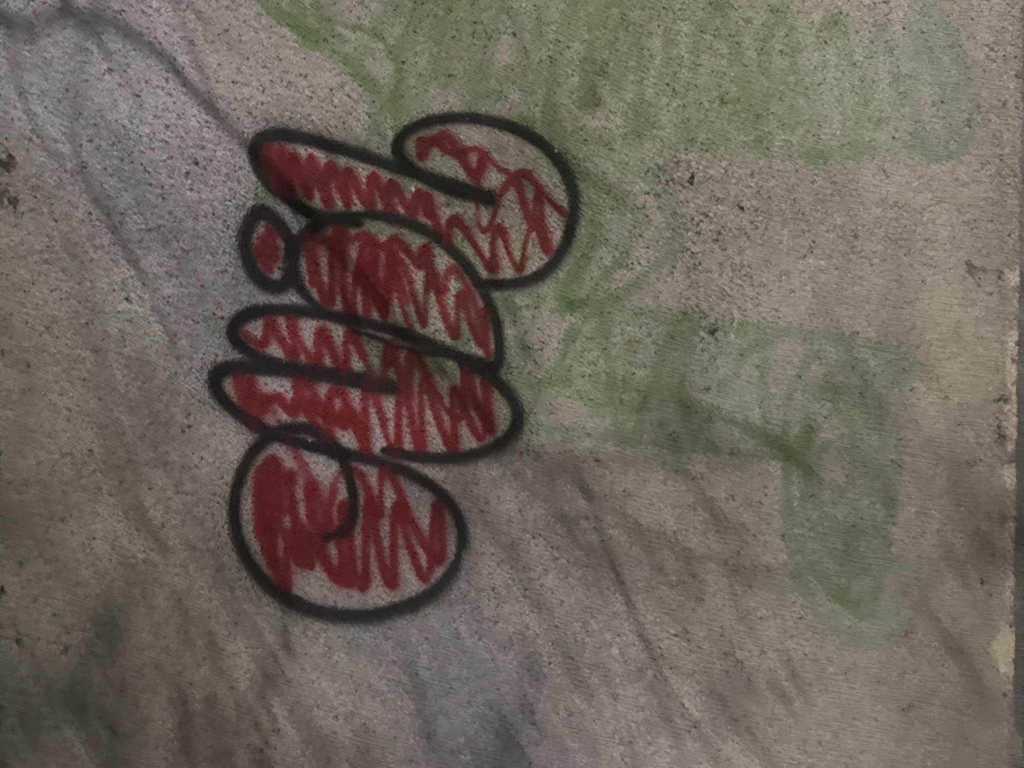 On a textured gray concrete surface, graffiti is visible. The graffiti is a stylized design composed of rounded shapes stacked vertically, outlined in black and filled with red scribble-like strokes. The design is positioned slightly off-center, toward the left side of the image. Green marks or smudges are visible on the right side of the concrete surface. No other people, objects, actions, or interactions are apparent. The location, Varna, Bulgaria, is not discernable from the image.