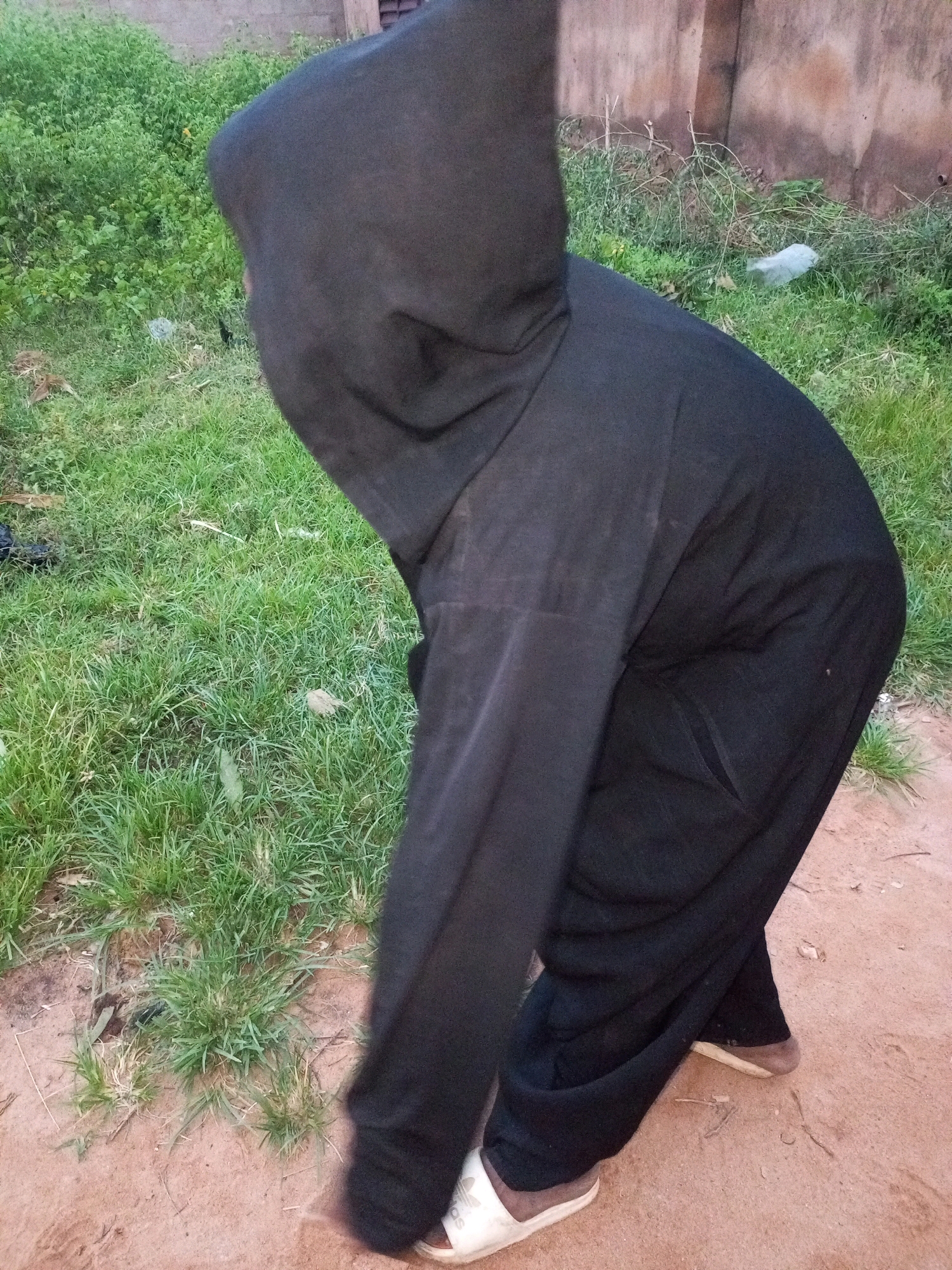 A person wearing a black hooded outfit and black pants is standing on a dirt path. The person's face is obscured by the hood. White sandals are visible on the person's feet. The background consists of green grass and a brown wall. The scene appears to be outdoors. The location is likely Tempure, Nigeria.