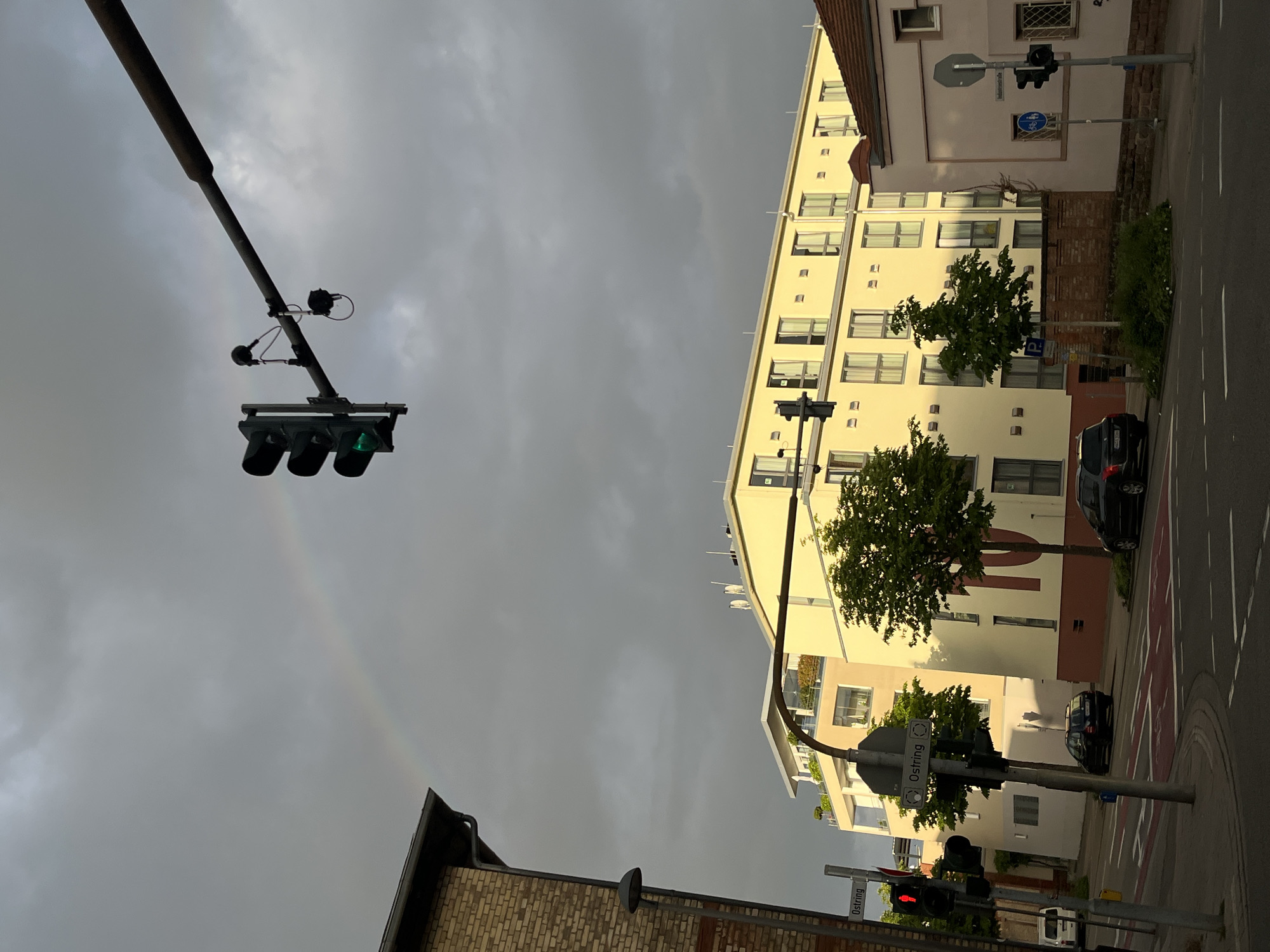 The image depicts a street scene in Landau in der Pfalz, Germany under a cloudy sky, with a faint rainbow visible. The perspective is tilted. Traffic lights are seen in the foreground, one showing a green light, the other a red light. Buildings line the street, including a light-colored apartment building. Cars are parked along the street. Street signs are visible, including one that reads "Ostring".