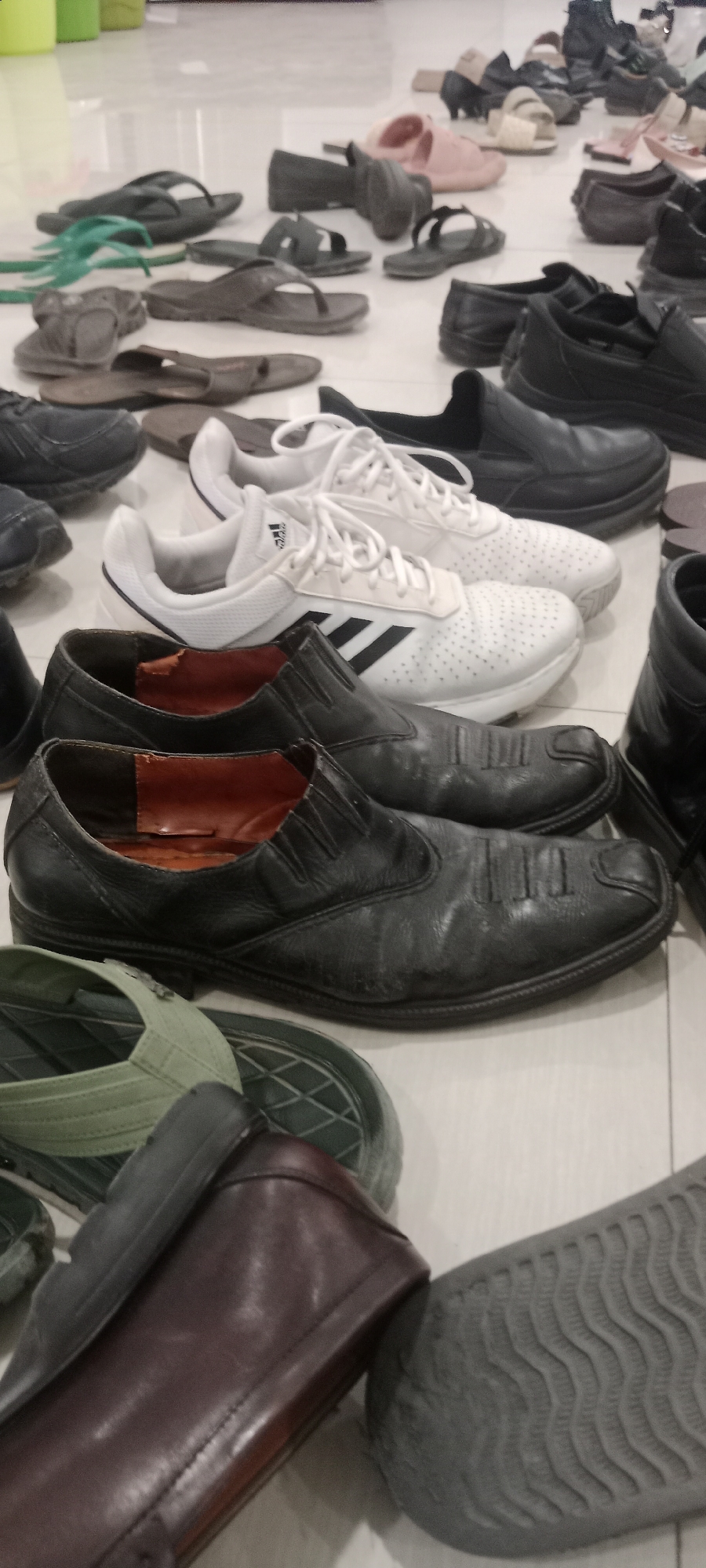 The image displays a collection of various footwear items scattered on a tiled floor, possibly in a public or communal area within Tuban, Indonesia. Among the footwear are black formal shoes, white athletic shoes with Adidas branding, and several pairs of sandals, including flip-flops and slides in shades of black, brown, and green. The arrangement suggests a casual or temporary placement rather than an organized display. The lighting appears to be overhead and diffused, casting subtle shadows on the floor and footwear. The overall scene depicts a utilitarian environment where shoes have been removed and left.