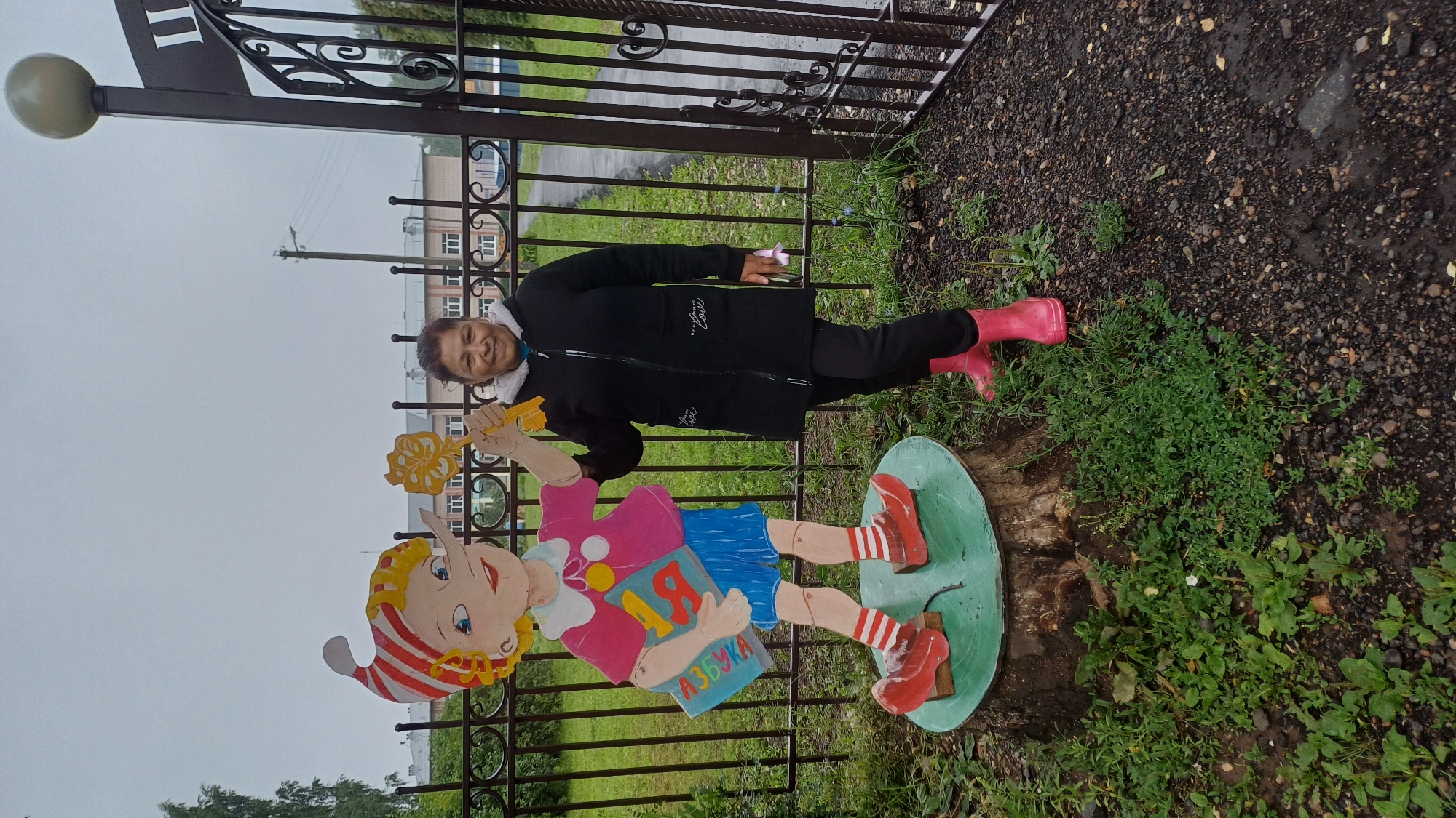 A woman stands next to a wooden cutout of Pinocchio in the city of Podgornoye, Russia. The woman is smiling and wearing a black jacket, black pants, and pink rain boots. She is holding a yellow, flower-shaped object in her right hand. The Pinocchio cutout is standing on a tree stump and is dressed in a pink shirt, blue shorts, and red shoes with white stripes. It holds a blue book with the word "Азбука" (Alphabet) written on it. The background shows a black metal fence and buildings in the distance under a cloudy sky. The ground is covered with gravel and some green plants.