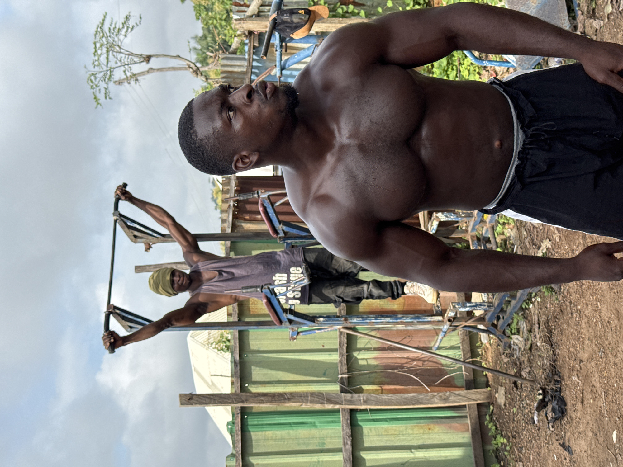 Two men are at an outdoor gym in Akwanga, Nigeria. The man in the foreground, facing right, is shirtless and wearing black shorts with white stripes. He has dark skin and a muscular physique. The man in the background, wearing a grey tank top and a green headwrap, is on a pull-up bar. He is also dark-skinned and appears to be mid-exercise. The gym equipment appears to be made of metal and is positioned on a dirt ground. The background shows a corrugated metal fence, some vegetation, and a cloudy sky.