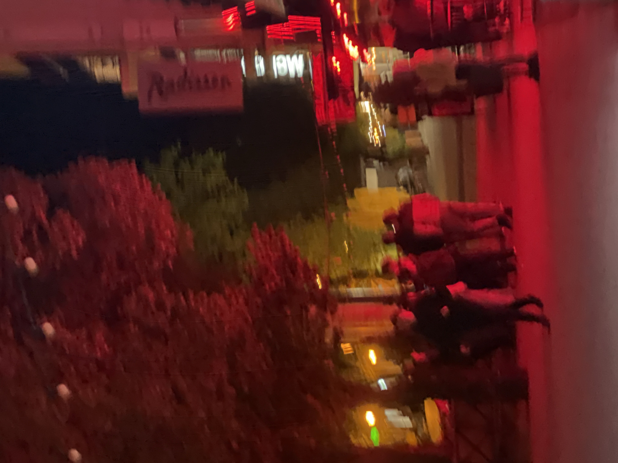 At night in Odesa, Ukraine, the Radisson Hotel is identifiable by its signage. The surrounding environment includes trees and streetlights. People are observed on the street.