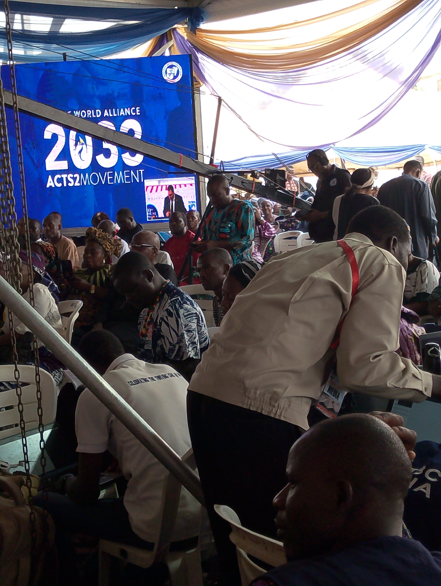 The image captures an indoor event taking place under a large tent or marquee in Dushepe, Nigeria, likely during daylight hours. The ceiling is adorned with draped fabrics in shades of blue, white, and brown/gold, supported by visible metal beams and chains. A large audience of adults is seated in rows of white plastic chairs, with many individuals visible from the back or side.
At the front of the gathering, a prominent blue LED screen displays the text "WORLD ALLIANCE," "2030," and "ACTS2MOVEMENT." A smaller inset screen on the main display shows a male speaker delivering a presentation. Among the attendees, a person is operating what appears to be a video camera on a tripod, indicating media coverage of the event. In the foreground, a man wearing a beige jacket and a red lanyard with a "MEDIA" badge is moving through the space.
Attendees are dressed in a variety of attire, ranging from casual wear to patterned traditional garments. On the back of a white shirt worn by a seated man, the text "CELEBRATING THE THREING OK" is partially legible. The visible text on the screen, along with the organized seating and media presence, suggests a formal conference, meeting, or public gathering centered around the "World Alliance 2030 Acts2Movement" initiative. The atmosphere appears attentive.