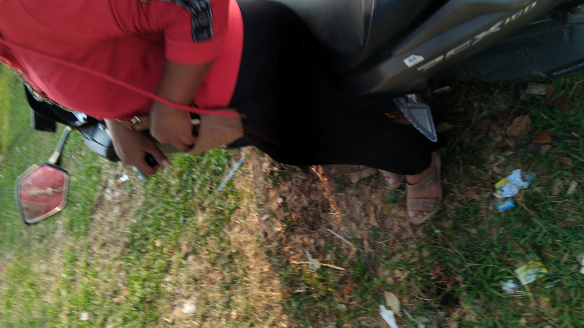 In Sembojo, Indonesia, a person is sitting on a motorcycle with their legs extended. They are wearing a red long-sleeved shirt and black pants, with brown sandals on their feet. The motorcycle is black and grey, with a silver trim on the side. The ground around the motorcycle is covered in grass and fallen leaves, with some litter scattered on the ground. A red mirror is visible on the left side of the motorcycle, and a small brown bag is hanging from the person's shoulder. The scene appears to be outdoors, possibly in a park or a roadside area.