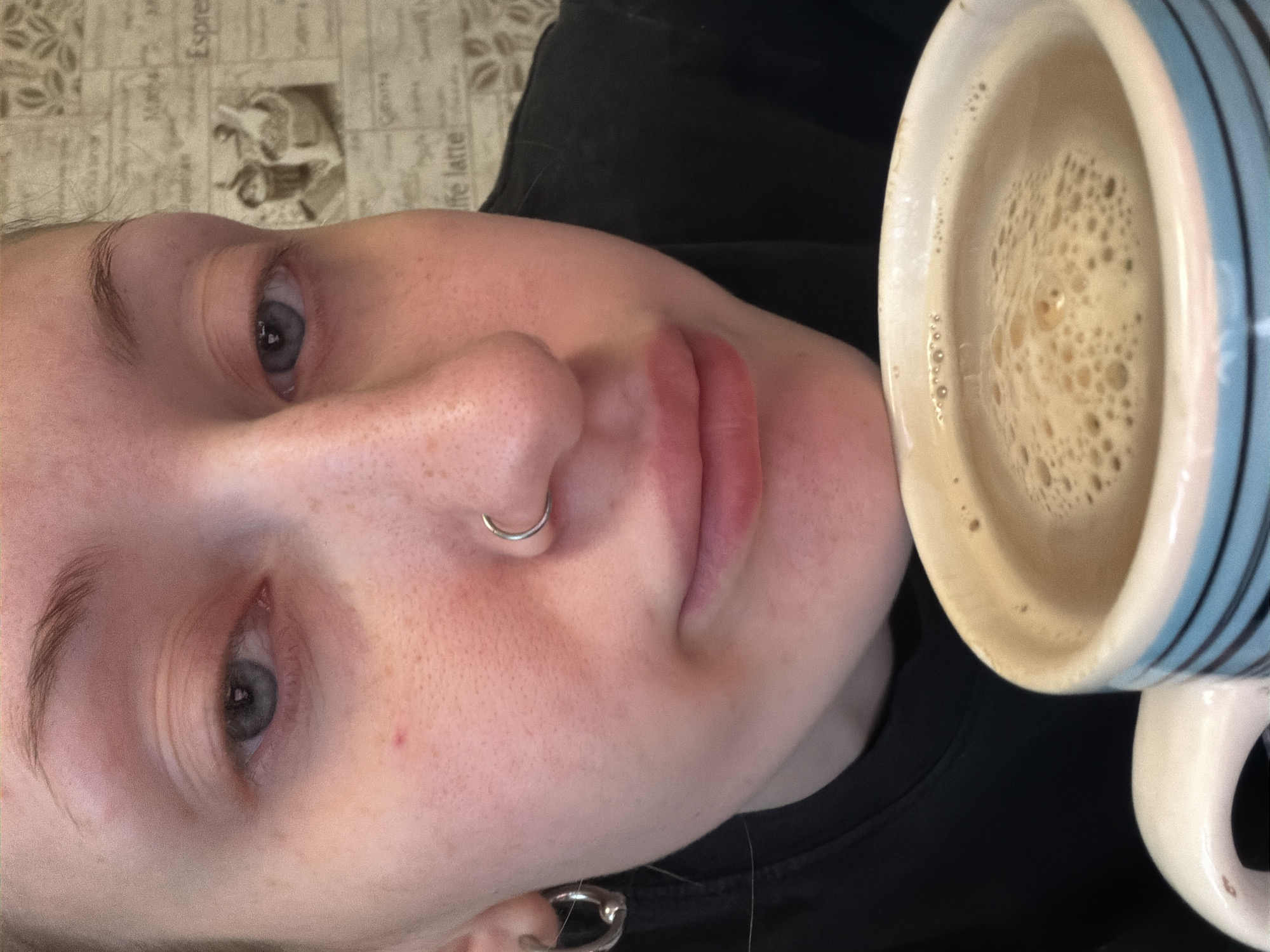 A close-up, slightly angled shot shows a person's face and a mug of coffee. The person has fair skin with freckles and a nose ring. Their eyes are a pale blue, and their lips are slightly parted. The background is a beige surface with a repeating pattern of text and images, possibly a placemat or tablecloth, with the words "Espresso" and "Cafe latte" visible. The coffee in the mug is light brown with a frothy layer on top, suggesting it's a cappuccino or latte. The overall mood is casual and relaxed, possibly indicating a morning routine or a break. The image is oriented sideways, and the lighting is soft, suggesting an indoor setting. The person appears to be looking towards the viewer, with a neutral or contemplative expression. There's no strong indication of time of day or weather. The location context mentions Saint Petersburg, Russia, but there are no specific visual cues to confirm this.