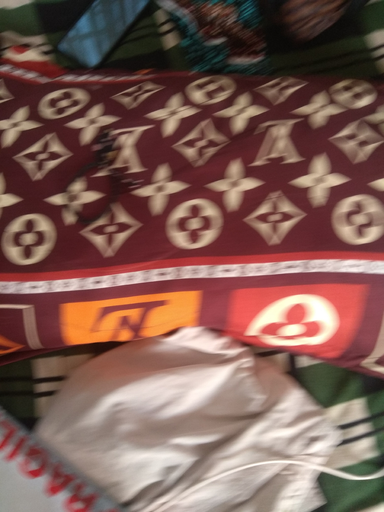 The image shows a close-up, blurry view of a red pillowcase with a Louis Vuitton-inspired pattern, featuring the brand's monogram and flower motifs in a light beige color. The pillowcase is folded and lies on top of other patterned fabrics, possibly bedding, with green, white, and black stripes visible. In the foreground, a crumpled, light-colored fabric object, likely a duvet cover or a pile of clothes, obscures the lower part of the pillowcase and some of the patterned bedding. The overall scene appears to be indoors, possibly a bedroom. No specific location in Jalingo, Nigeria, can be determined from the visual information provided.