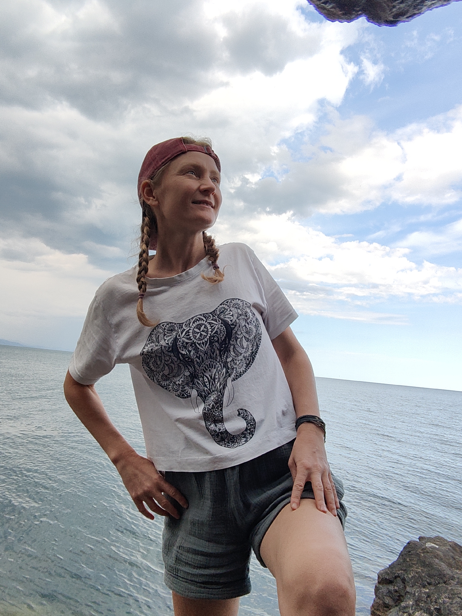 A woman stands by the sea with her hands on her hips, looking up and to the right, with a contented expression. She wears a white t-shirt with an elephant design, a red baseball cap, and dark shorts. Her blonde hair is styled in two braids. The sea is calm and stretches to the horizon under a cloudy sky. The location appears to be the coast of Utios, Ukraine.