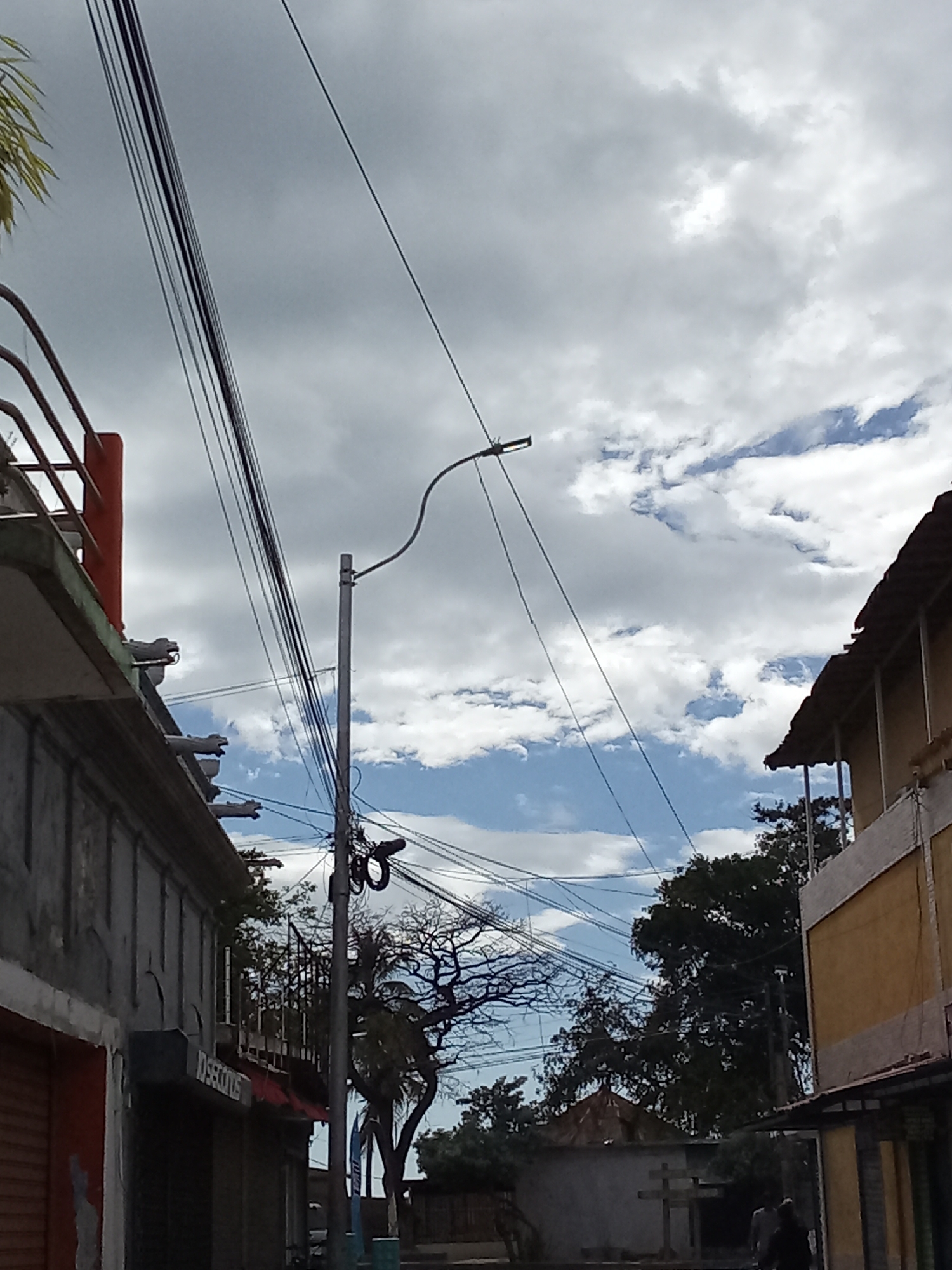 The image shows a street scene in Juan Griego, Venezuela. Utility poles and wires crisscross the sky, partially obscuring a cloudy sky with patches of blue. On the left is a multi-story building with a faded gray facade, a balcony with a red awning, and decorative gargoyles along the roofline. The word "DISECTONDS" is visible on a sign. On the right is another multi-story building with a yellow facade and a dark roof. In the background, between the buildings, are trees, a small structure with a pointed roof, and a wooden cross. Two people are walking in the lower right corner of the image.