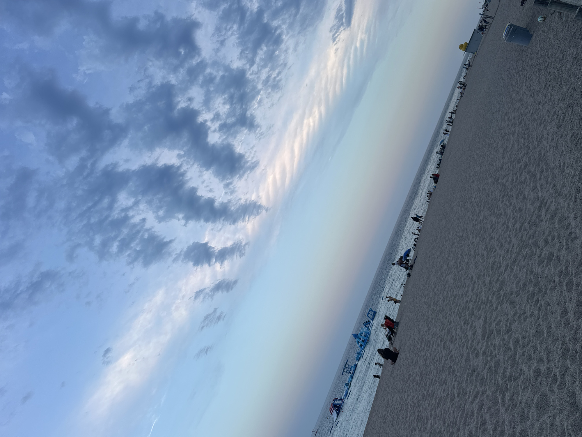 The image depicts a beach scene with a diagonal composition, tilting upwards from the bottom right to the top left. The foreground is dominated by sand, textured with numerous small ripples. In the middle ground, a body of water, likely the sea, stretches horizontally across the frame. Several inflatable water park structures are visible on the water, appearing as colorful, modular obstacles. Scattered across the sand are numerous people in various states of repose and activity. Some are sitting or lying on beach chairs and towels, while others are standing or walking. A few individuals are in the water near the inflatable structures. The background is composed of the sky, which transitions from a pale blue at the horizon to darker, cloud-filled hues at the top. The clouds are varied, with some appearing wispy and others more dense and textured. The lighting suggests it is either early morning or late afternoon, casting a soft, diffused light over the scene. The specific location is not identifiable from the image itself, and therefore cannot be confirmed as Blanes, Spain.