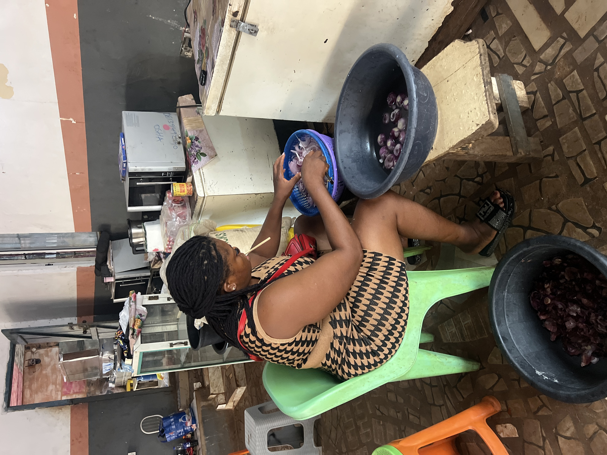 In Nkpor, Nigeria, a woman with braided hair, wearing a patterned dress, is seated on a green plastic chair in a cluttered room. She is actively slicing onions, placing them into a blue plastic basket. A dark basin filled with sliced onions in water sits on a wooden stool beside her, and another large black basin containing chopped onions is on the floor. The background reveals a busy environment with appliances like a microwave, a refrigerator, and a display case, suggesting a commercial or market setting. The walls are painted in distinct horizontal bands of grey and beige, with some areas showing wear and tear. Several plastic chairs and containers are scattered around the room, contributing to the overall impression of a working space.