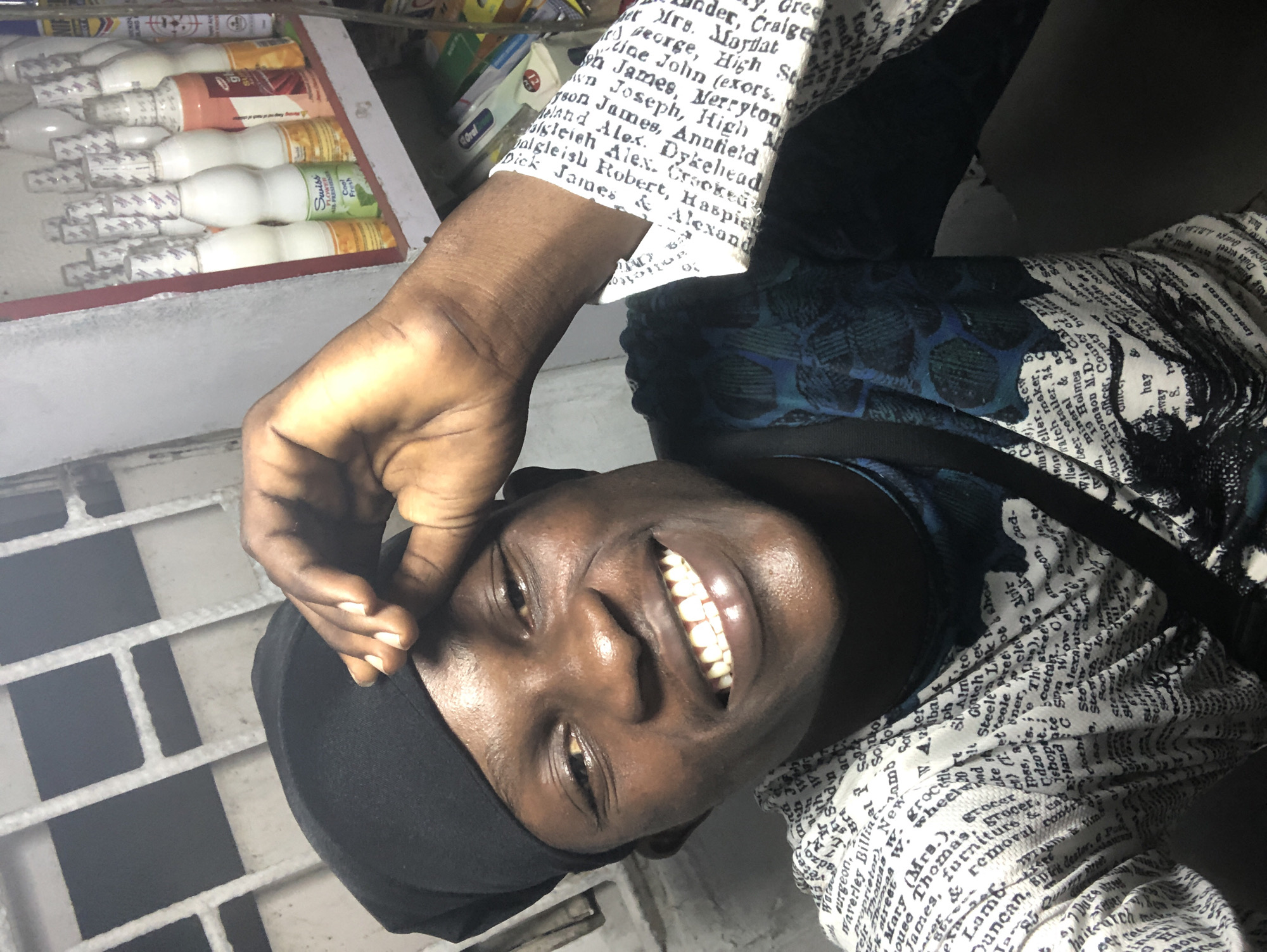 The image is a close-up, selfie-style portrait of a young Black man wearing a black durag and a white shirt with a text print. He is smiling broadly with his mouth open, revealing his teeth. His right hand is raised to his forehead, touching the durag. Behind him, on the right side of the frame, a shelf is visible filled with various bottles, some of which appear to be cosmetic or health products. One bottle in the foreground has a label that says "Swiss Flower Air Freshener Cool Fresh". The background on the left shows a metal grate structure, possibly part of a window or ventilation. The overall lighting suggests an indoor setting, possibly a shop or market stall. The text print on the man's shirt is a dense arrangement of words, appearing to be from an old newspaper or book. The setting is consistent with a retail environment in Ado, Nigeria, given the presence of common consumer goods and the urban backdrop.