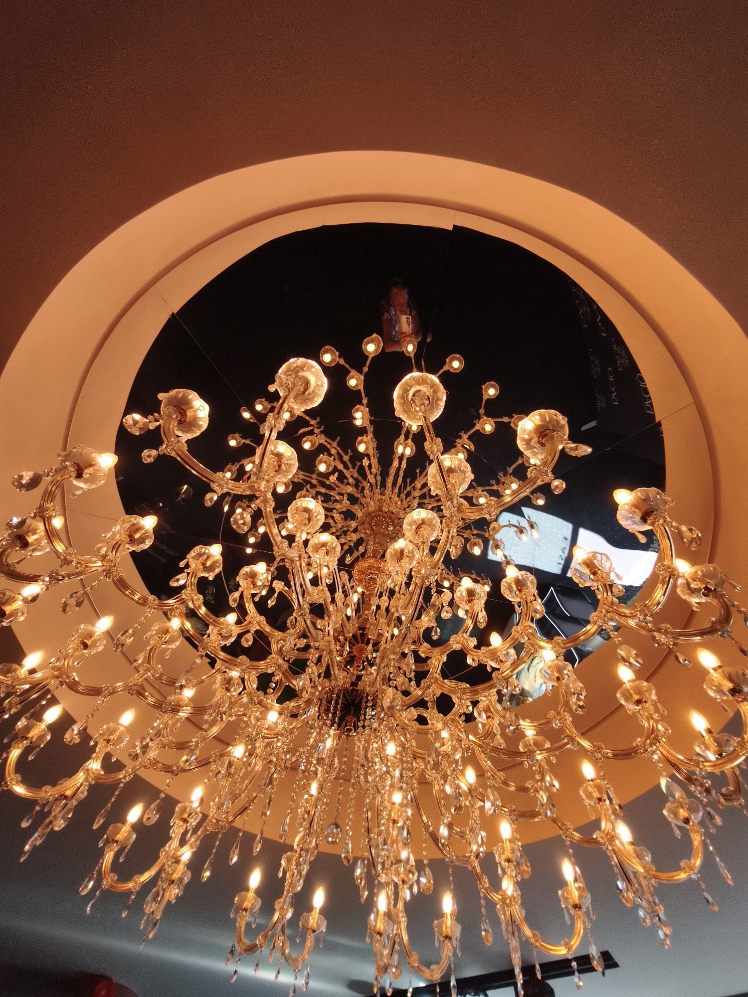 An ornate, golden chandelier hangs from a ceiling in a circular opening. The chandelier features numerous crystal droplets and electric candles. The ceiling is a warm, peach color, and the circular opening reveals a dark, reflective surface, possibly a mirror or a dark ceiling, with faint reflections of lights and a person reading. The overall impression is one of opulence and dramatic lighting. The specific location in Tallinn, Estonia, is not identifiable from the image.