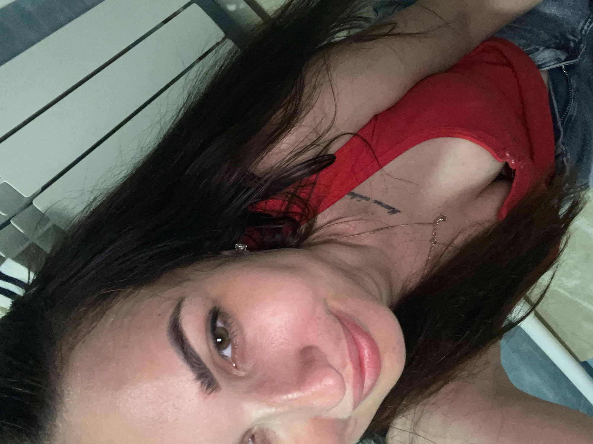 The image is a close-up, waist-up selfie of a woman with dark hair and brown eyes, wearing a red tank top and blue jeans. She has a tattoo on her chest and is wearing a necklace. Her hair is partially covering her face and shoulder. The background appears to be indoors, with a white radiator visible on the right side of the frame. The lighting suggests it may be daytime. The scene appears to be a personal setting, possibly a bedroom or living area. There is no indication of the city or location.