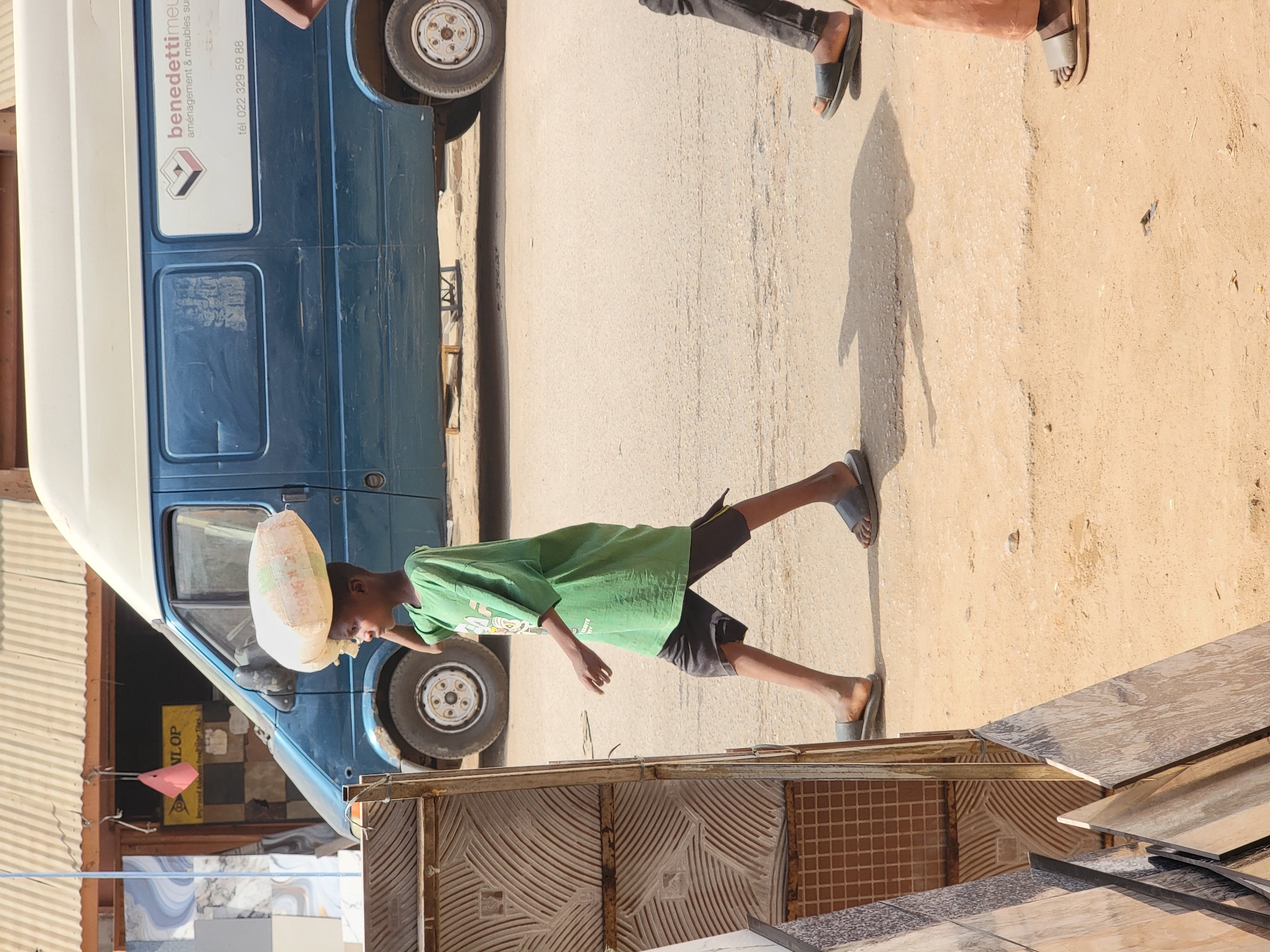 A boy in a green t-shirt carries a large, white sack on his head while walking on a dirt road next to a blue van. The scene occurs in Maiburiji, Nigeria. Other people are nearby, and are wearing sandals. In the foreground there are building materials for constructing buildings.