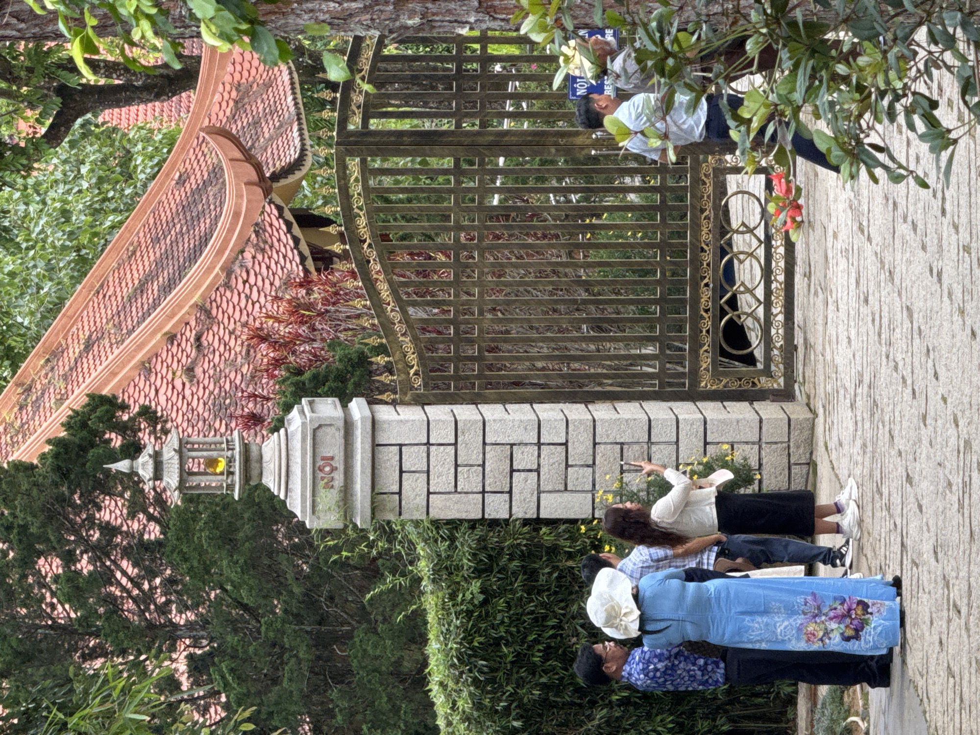 The image captures an outdoor scene at the entrance of a Vietnamese establishment, possibly a temple or a historical site, located in Da Lat, Vietnam. The architecture features a traditional tiled roof with curved edges, and a tall stone pillar adorned with a decorative lantern and engraved text, reading "NÔI" in Vietnamese. A decorative metal gate with intricate patterns stands to the right of the pillar, serving as an entrance.
In the foreground, a group of four individuals are gathered on a stone-paved pathway. One person, a woman wearing a long blue dress with floral patterns, faces away from the camera, while a woman in a white hat and a man in a blue patterned shirt stand nearby. A woman with long dark hair is holding a smartphone, seemingly taking a picture or recording a video. Two men are visible in the background, near the gate. One is wearing a light blue shirt and dark pants, and the other is wearing a white shirt and dark pants. The surrounding environment is lush with greenery, including dense bushes and trees, suggesting a garden or park setting. The overall atmosphere appears serene and possibly tourist-oriented, given the presence of individuals engaged with their surroundings and capturing memories.