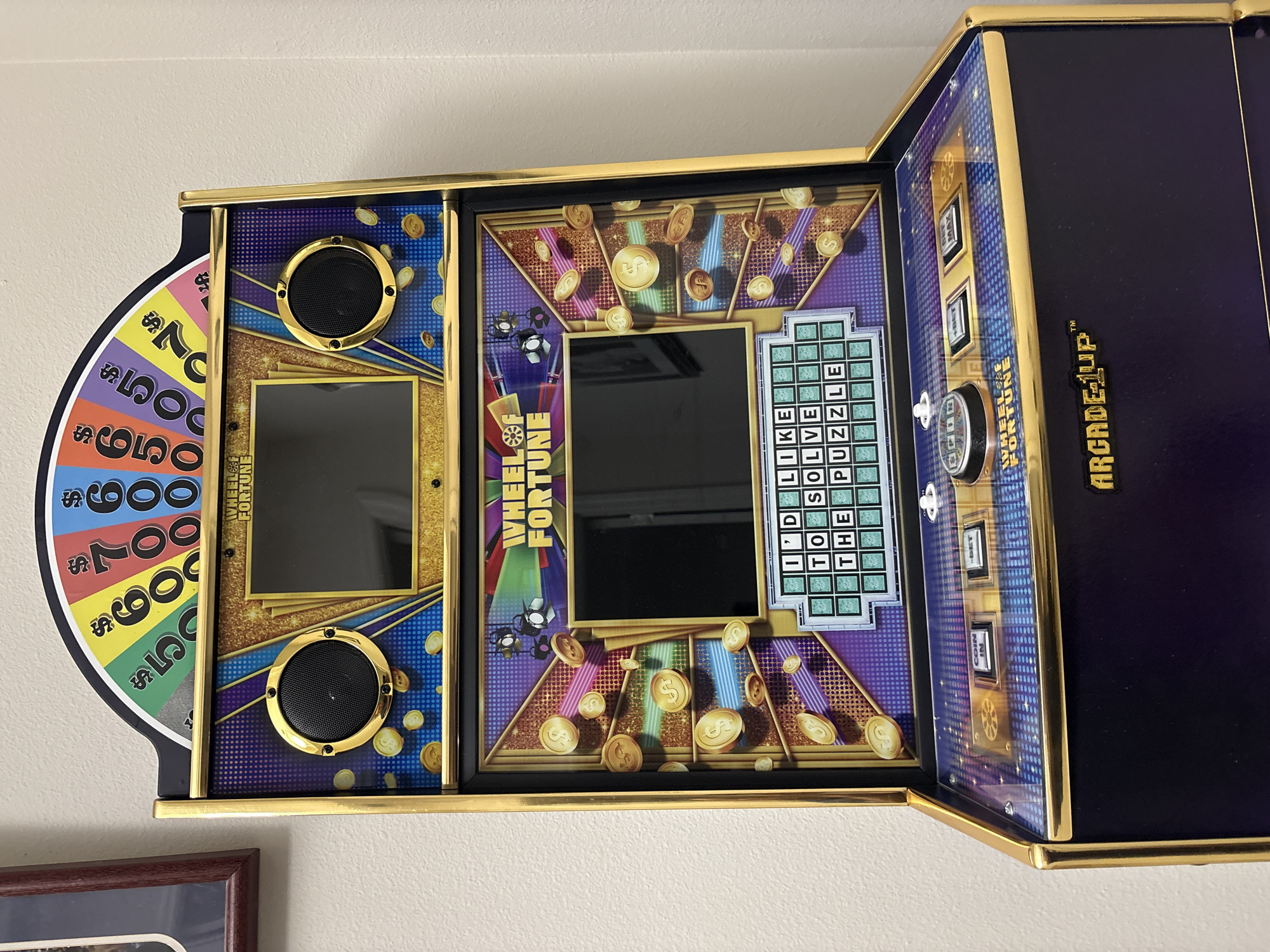 The image shows a close-up, eye-level, frontal view of a Wheel of Fortune arcade game machine, predominantly black and gold, with a curved top displaying a colorful spinning wheel with dollar amounts. The machine features two screens: an upper screen with the "WHEEL OF FORTUNE" logo and speakers on either side, and a lower screen that shows a puzzle board with the text "I'D LIKE TO SOLVE THE PUZZLE." Gold coins with dollar signs are depicted as graphics scattered across the machine's surface. Below the lower screen are control buttons, including "SPIN," "BET 1," and "BET MAX," and the "ARCADE1UP" logo. The machine is positioned indoors, against a plain, lightly textured off-white wall. A portion of a framed picture with a dark brown frame is visible on the left edge of the image. The scene appears to be in a home or arcade setting, and no specific city or location is discernible from the visual information.