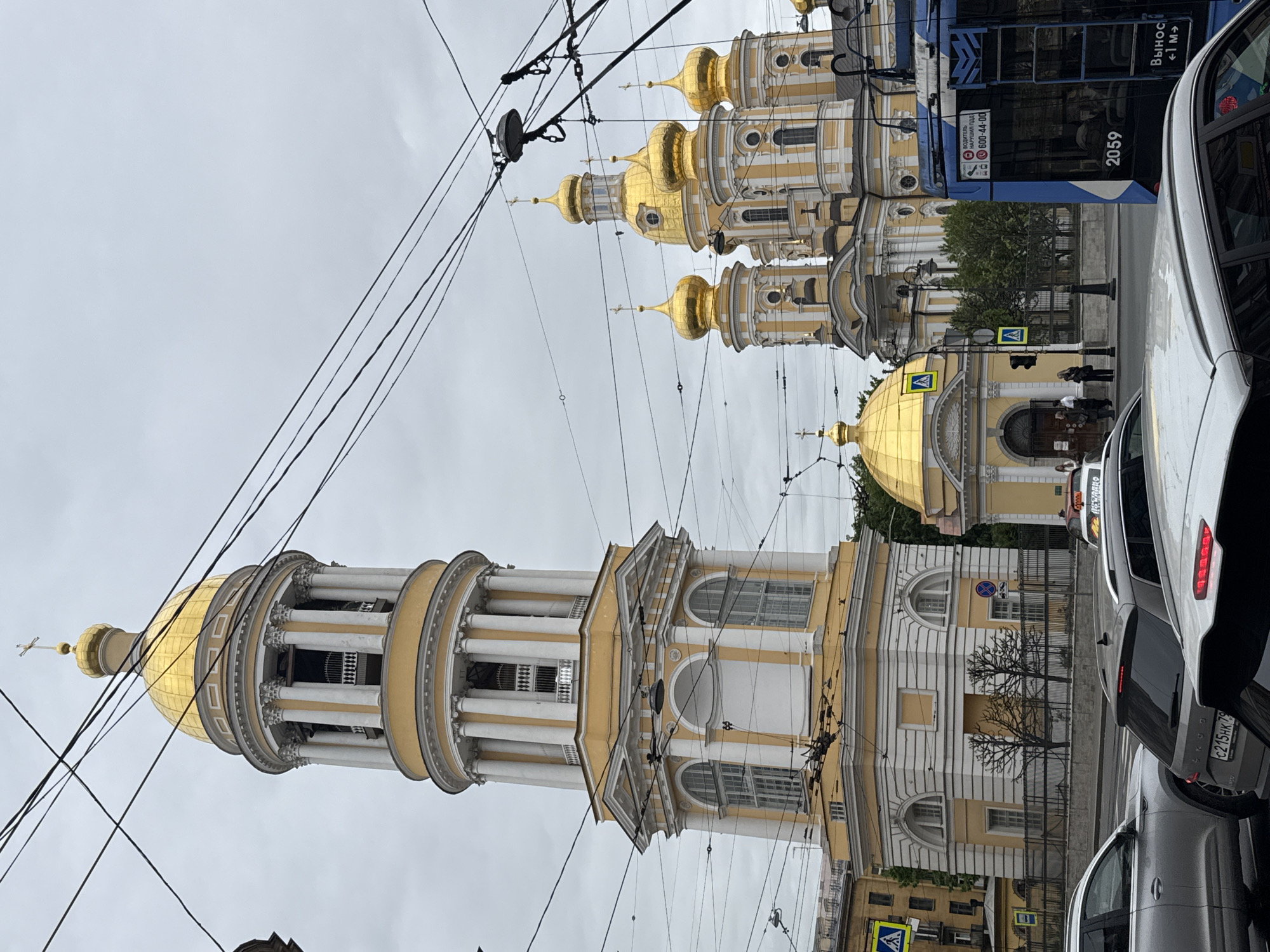 The image captures a street scene in Saint Petersburg, Russia. The focal point is the Smolny Cathedral, characterized by its gold domes and white and yellow façade. Power lines crisscross the sky, adding a complex network to the urban landscape. The scene includes modern vehicles: a blue trolleybus and several cars, indicating contemporary city life. People are seen walking near a pedestrian crossing, and buildings are visible along the street, reflecting the architectural style of Saint Petersburg.