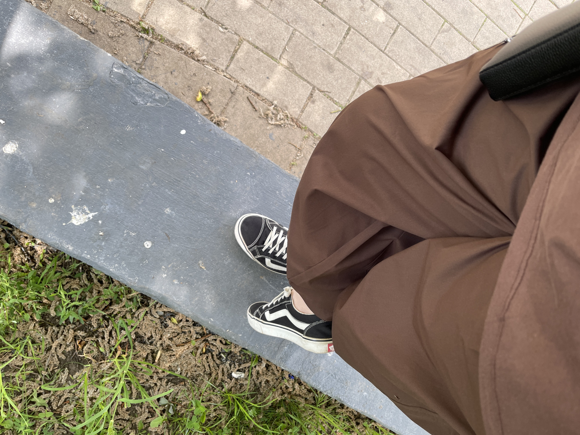 The image shows a person sitting on a gray bench. They are wearing brown, loose-fitting pants and black and white sneakers. A black bag is partially visible next to their leg. The bench is located next to a patch of grass and debris. In the background, there is a brick-paved area. The image provides no information about the location in Moscow, Russia.