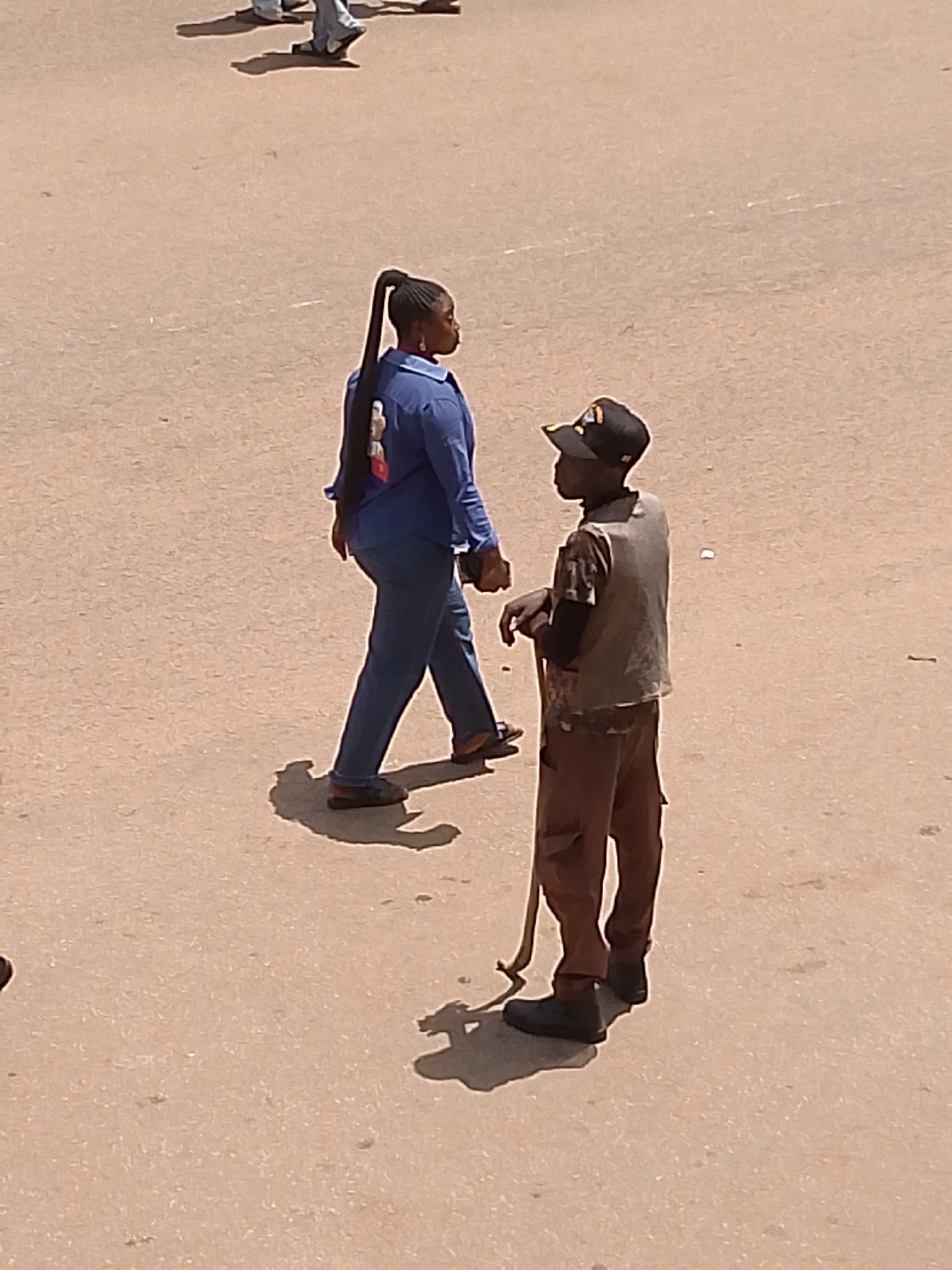 In Bukuru, Nigeria, a woman with a long black ponytail is walking away from the camera. She is wearing a blue long-sleeved shirt and blue pants. Beside her, a young person in a brown baseball cap and a light-colored vest over a patterned shirt and brown pants stands holding a long stick. The ground is a dusty, sandy surface. In the upper left corner, the blurred legs and feet of other people are visible.