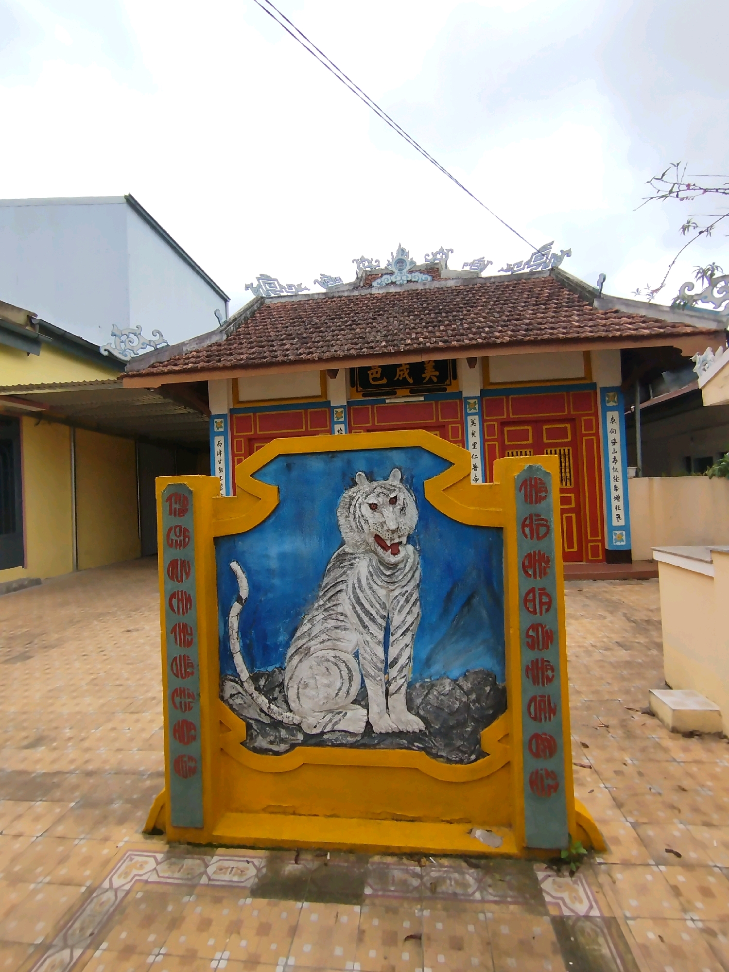 A static, medium-shot photograph captures a vibrantly painted yellow and blue shrine featuring a white tiger. The tiger, depicted with a fierce expression and red eyes, is seated on a rock in front of a blue background. The shrine itself is adorned with red lettering on a light blue vertical panel on its sides. Behind the shrine, a building with a red facade, yellow trim, and a tiled brown roof is visible. The building's roof has ornate, decorative elements. To the left of this building, a yellow structure with a silver awning is partially visible. The ground is paved with light brown tiles, some of which are decorated with a white and darker brown patterned border. The overall scene appears to be outdoors, likely in a temple or a place of worship, given the architectural style and the presence of the shrine. The sky is overcast and bright. The image does not contain any people or animals other than the painted tiger. Based on the architectural style, it is likely located in Da Lat, Vietnam, a city known for its French colonial and Vietnamese-inspired architecture.