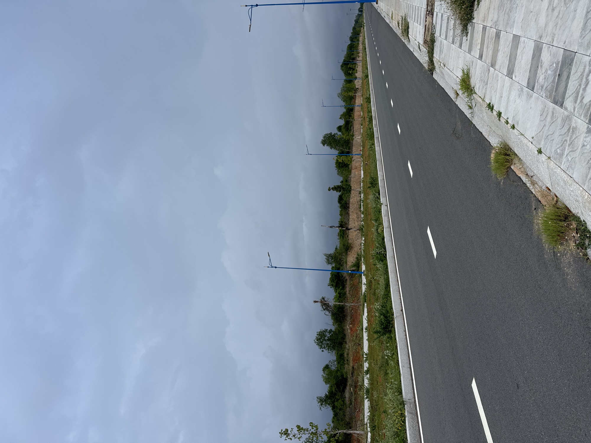 The image depicts an asphalt road with white lane markings and a sidewalk paved with large, light grey and white square tiles. The road is lined with sparse green vegetation and several tall, blue streetlights. In the background, there is a low stone wall and a line of trees under a cloudy, overcast sky. The scene appears to be an undeveloped area or a newly constructed road in Mui Ne, Vietnam, possibly a coastal town known for its beaches and sand dunes. There are no people or vehicles visible in the image. The overall impression is one of emptiness and potential.