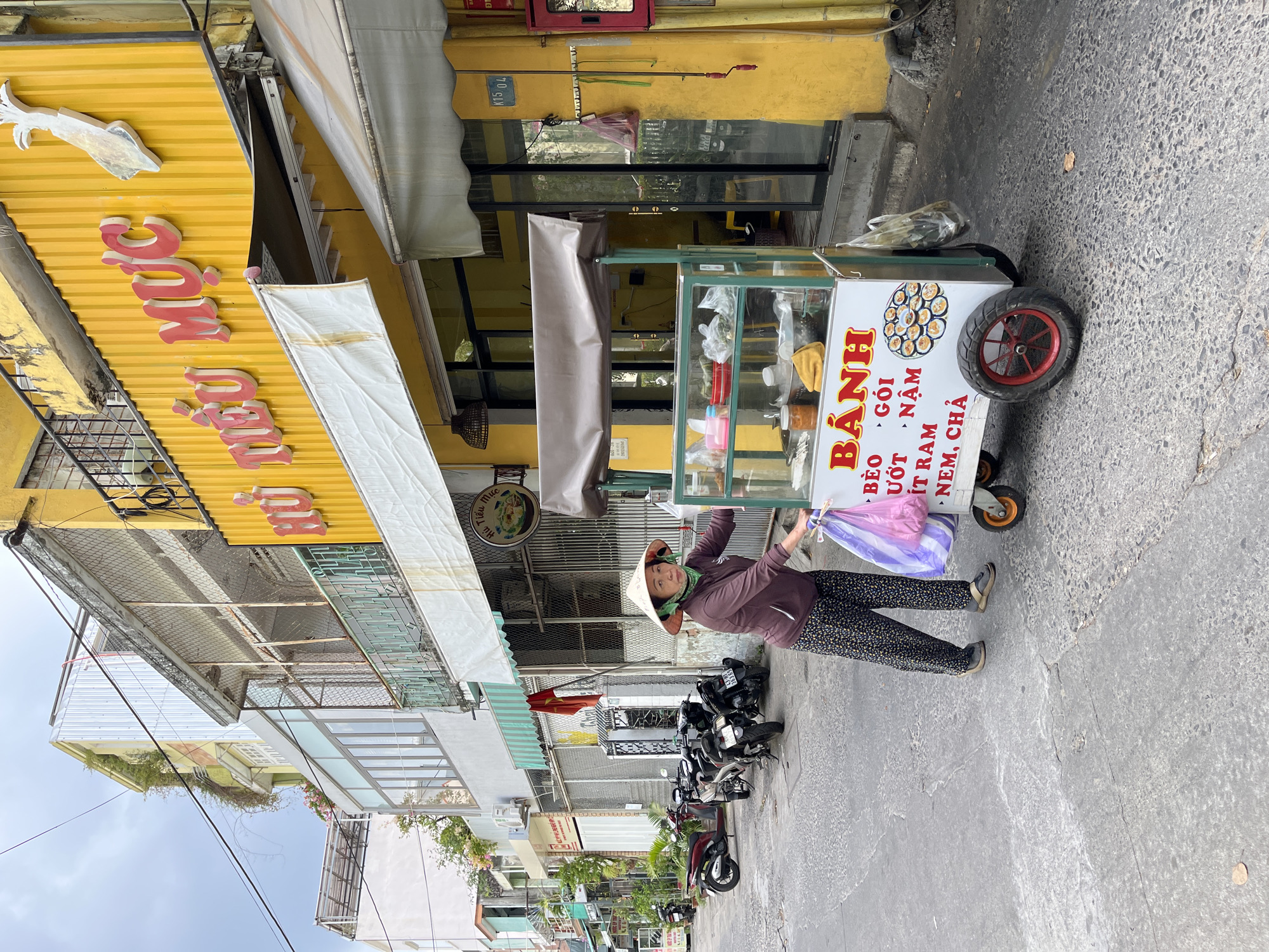 An outdoor street scene in Đà Nẵng, Vietnam, captures a woman vendor with her food cart on what appears to be an overcast day. The image is taken from a slightly elevated perspective, looking down a bustling narrow street or sidewalk.
The woman, wearing a traditional Vietnamese conical hat (nón lá), a dark long-sleeved top, and patterned trousers, stands on the right side of her mobile food cart. She holds a plastic bag, colored purple and clear, in her left hand, looking towards the camera with a neutral expression.
The white food cart features a glass display case showcasing various ingredients and containers, indicating different types of Vietnamese dishes. Prominent red Vietnamese text on the cart reads "BÁNH" (cake/dumpling), with specific dish names listed below: "BÈO", "GÓI", "ƯỚT", "ÍT RAM", "NẬM", and "NEM, CHẢ". An illustration of small round dishes is also visible on the cart's side.
Behind the woman and the cart, several motorbikes are parked along the street. The background is dominated by yellow-painted buildings with awnings. One striking yellow building features large red Vietnamese letters spelling "Mực" (Squid), along with smaller text "HỦ TIẾU MỰC" (Squid Noodle Soup) and "K15 04". The sky above is cloudy, suggesting daytime. The street surface is paved with asphalt or concrete. The scene offers a glimpse into typical daily street life and the vibrant street food culture of Vietnam.