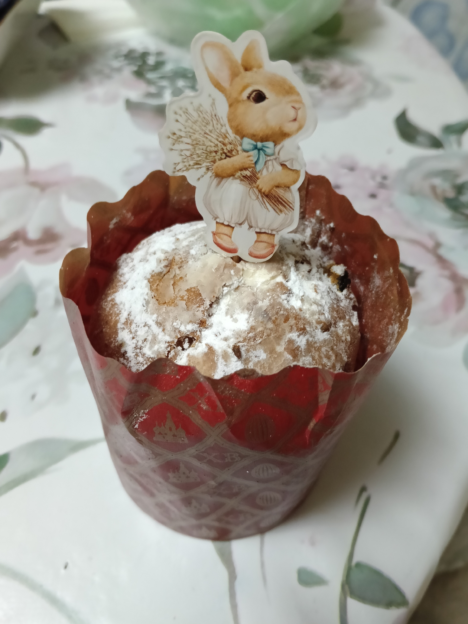 A close-up shot of a cupcake is placed on a floral-patterned surface. The cupcake is in a red paper wrapper with a geometric pattern. A rabbit-shaped paper decoration is stuck on top of the cupcake, which is covered in powdered sugar. The location is unknown.