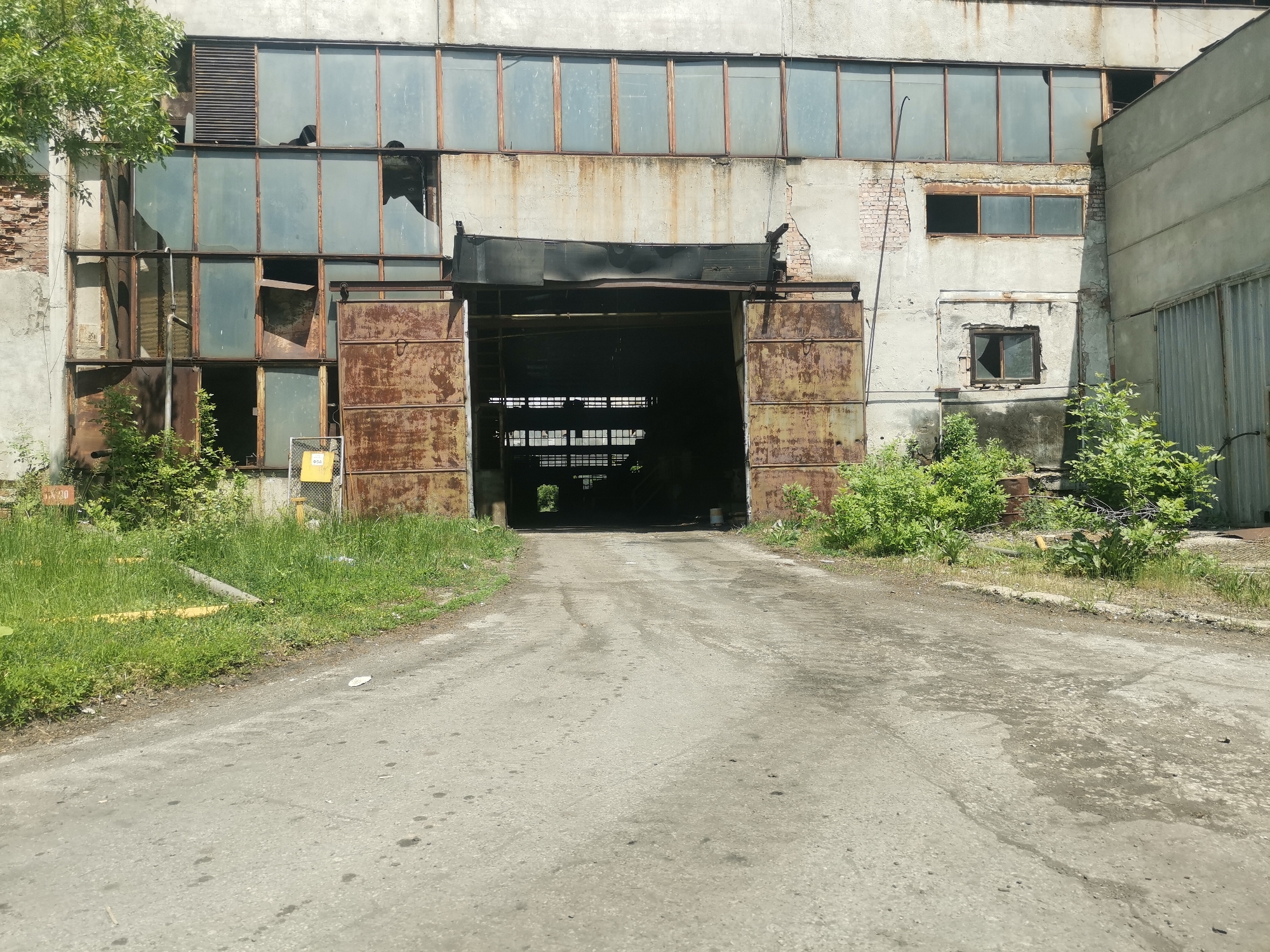 The image depicts an abandoned industrial building, possibly a warehouse or factory, in Zhivkovo, Bulgaria. The building's facade is dilapidated, with broken windows, crumbling brick, and rusty metal doors that are open, revealing a dark interior. Vegetation is growing along the base of the building, and a cracked asphalt driveway leads towards the entrance. There are no people visible in the frame. The overall impression is one of decay and disuse.