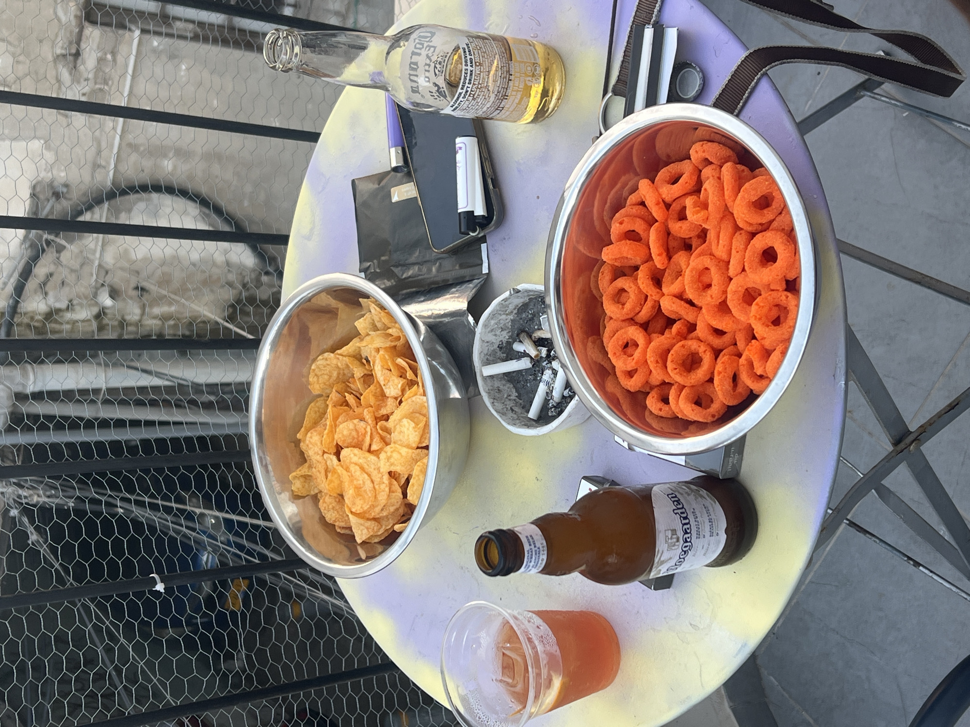An outdoor scene features a circular table on a patio in Tel Aviv, Israel. The table is laden with snacks and drinks. Two metal bowls are present: one filled with potato chips and another with cheese puff rings. A plastic cup contains a reddish beverage with ice and a slice of lemon. Two beer bottles are on the table: a clear glass bottle with a yellow label, and a brown glass bottle with a white label. An ashtray filled with cigarette butts sits between the bowls. A smartphone and two lighters are also on the table. A metal fence with a hexagonal mesh pattern forms the backdrop, with a glimpse of a building and a bicycle wheel visible behind it. The overall impression is one of a casual gathering or relaxation.