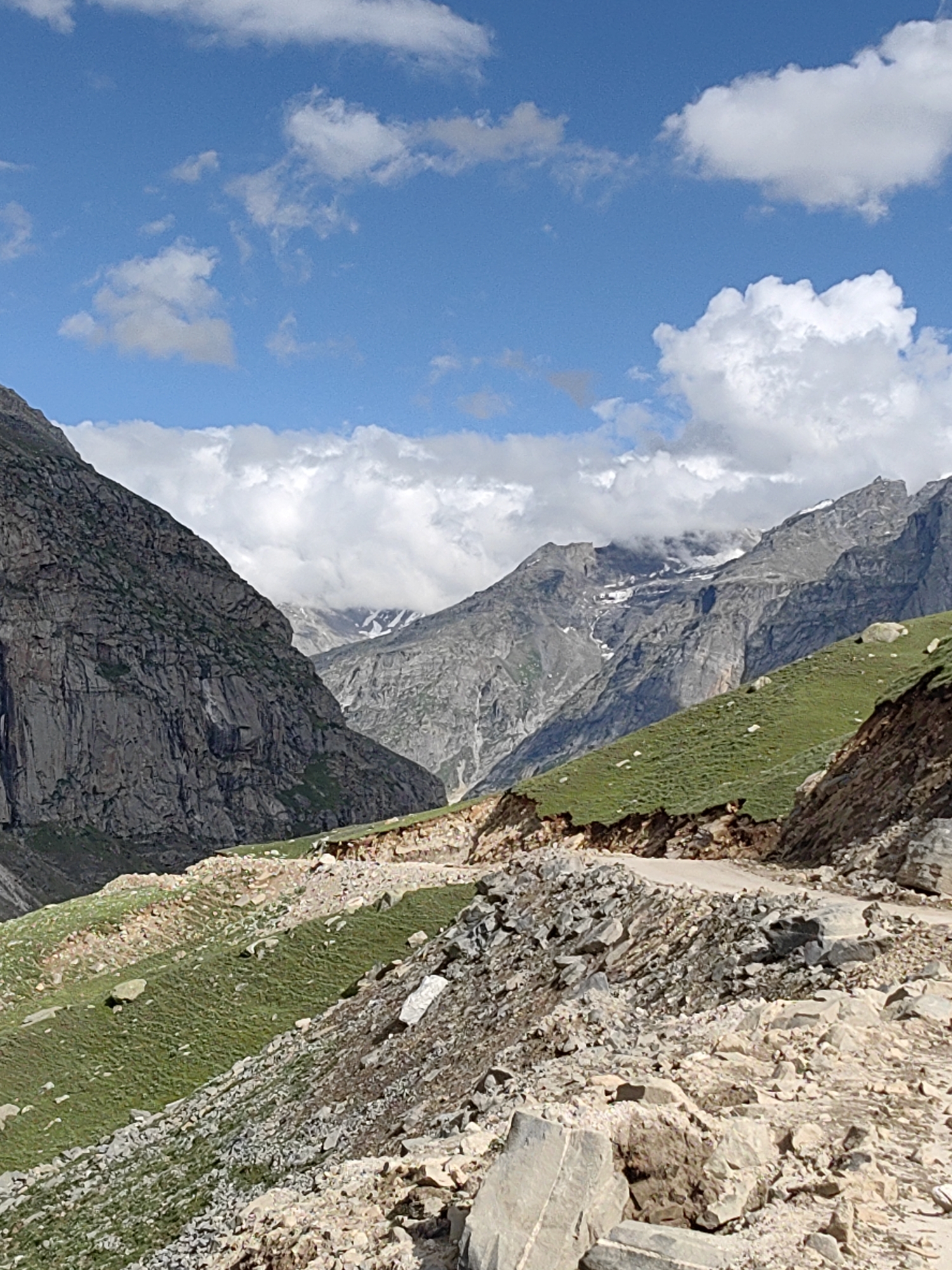 The image shows a mountain landscape with a road winding through it. The terrain is rugged and rocky, with steep slopes covered in patches of green grass. Tall, imposing mountains rise in the background, some with snow-capped peaks. The sky is blue with fluffy white clouds. There are no people or animals visible in the image. The scene suggests a remote, high-altitude location, consistent with the mountainous regions of India.