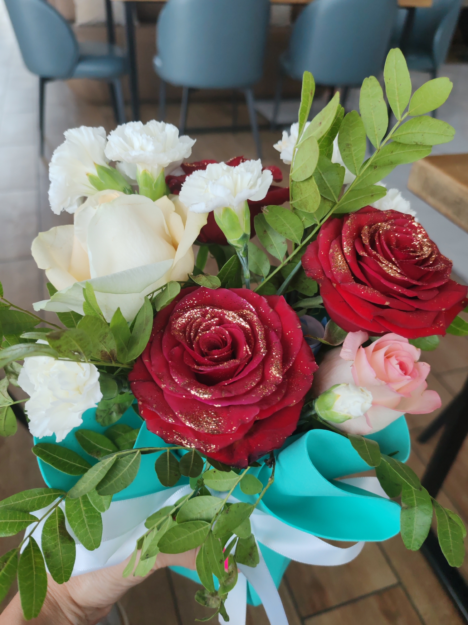 A close-up, eye-level shot focuses on a bouquet of flowers, held by a person's hand which is partially visible on the left side of the frame. The bouquet is arranged in a bright teal gift box adorned with a white ribbon. The flowers include several deep red roses with gold glitter accents, a creamy white rose, and white carnations. Green foliage from the bouquet and some decorative branches with small leaves frame the flowers. The background is blurred, showing the interior of what appears to be a cafe or restaurant, with grey chairs and tables. The scene is brightly lit. The exact location within Amursk, Russia is not determinable from the image itself.