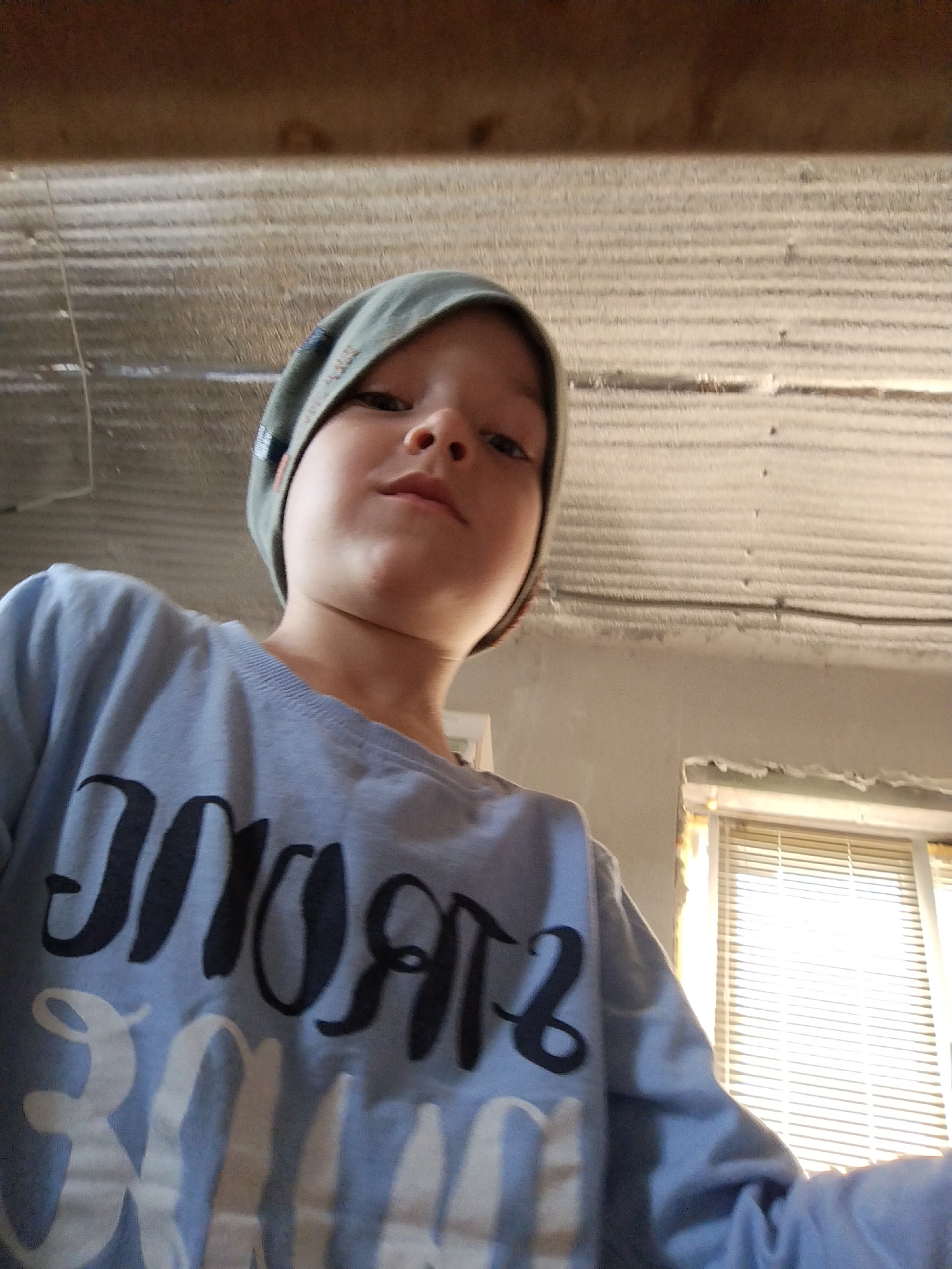 A low-angle, waist-up shot shows a young child wearing a light blue long-sleeved shirt and a green beanie. The child is looking upwards towards the camera, with their expression neutral. The shirt has large, stylized black and white text printed on it, which appears to read "STRONG".
The background is utilitarian. Above the child, there is a corrugated metal ceiling, possibly made of aluminum, with visible wires running across it. Below the ceiling, a grey concrete wall is visible. To the right, a window with white horizontal blinds is letting in bright daylight, suggesting it is daytime. The overall scene appears to be indoors, in a simple, perhaps unfinished or industrial-looking space.
The context provided indicates the location is Zavodoukovsk, Russia. The lighting suggests it is daytime. There are no obvious weather cues. The emotions conveyed are difficult to ascertain precisely due to the low angle and neutral expression, but the overall impression is one of straightforward observation.