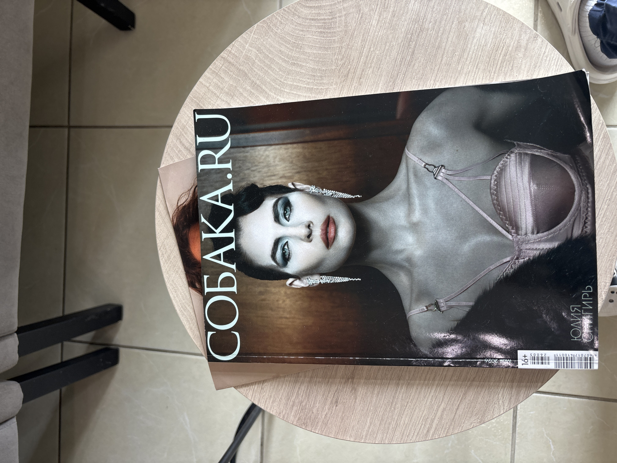 A magazine titled "СОБАКА.RU" with a woman in a bra on the cover is resting on a light-colored round table. The magazine cover features a woman with dark hair and bright makeup wearing a bra and long, sparkly earrings. The table is positioned on a tile floor, and a piece of furniture is partially visible on the left side of the image. The location is Vsevolozhsk, Russia.