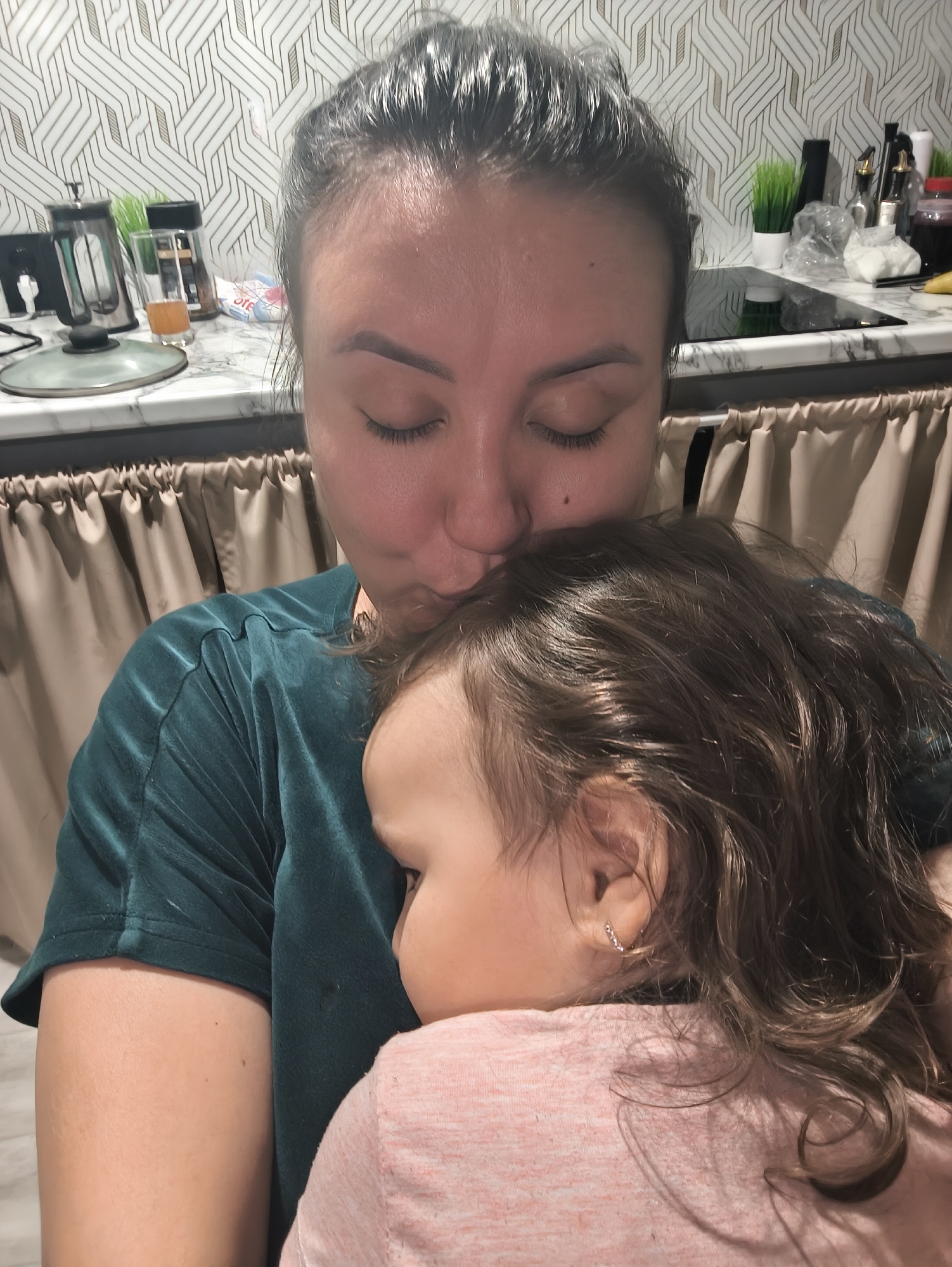 A woman with dark hair pulled back, wearing a dark green velvet-like top, is kissing the top of the head of a young child with dark curly hair and an earring, who is wearing a light pink shirt. They are indoors, with a kitchen in the background. The kitchen features a counter with a marble-like surface and various appliances and kitchenware, including a french press, jars, and bottles. The backsplash has a geometric pattern. The curtains are light brown. This photo does not contain locational information of Сокуры, Russia.
